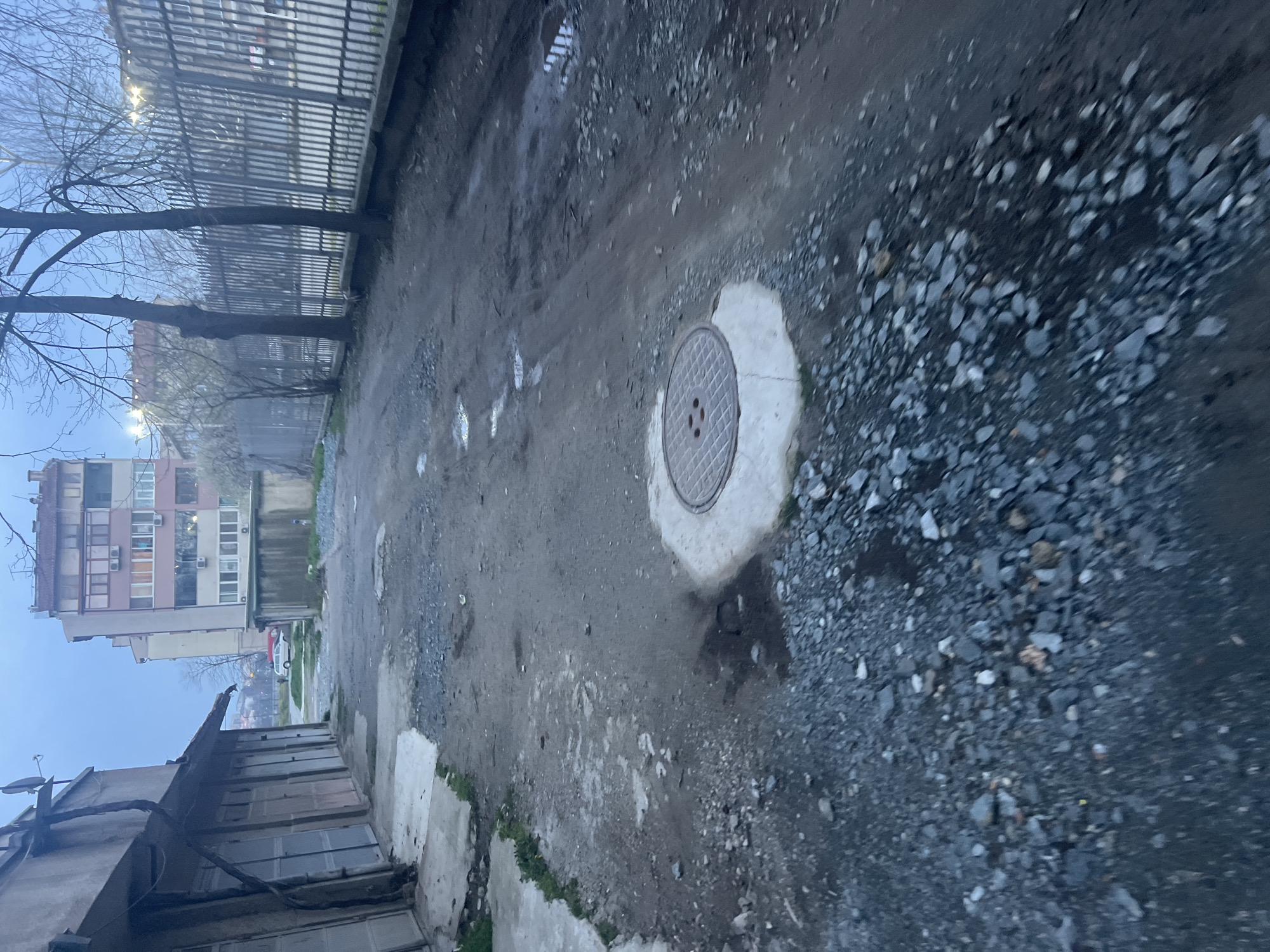 The image depicts a view of an unpaved alleyway or side street in a residential area. The ground is a mix of dirt, mud, puddles, and gravel. A manhole cover sits in the middle of the alley. To the left, there's a weathered, low-rise building, and in the distance, a taller apartment building is visible. A fence runs along the alley on one side. Bare trees stand along the fence line. The sky appears overcast. Given the presence of apartment buildings and a generally urban environment, it is likely this scene is located in a city; based on context provided, the location is likely the city of Yambol, Bulgaria. No people are visible in the image.
