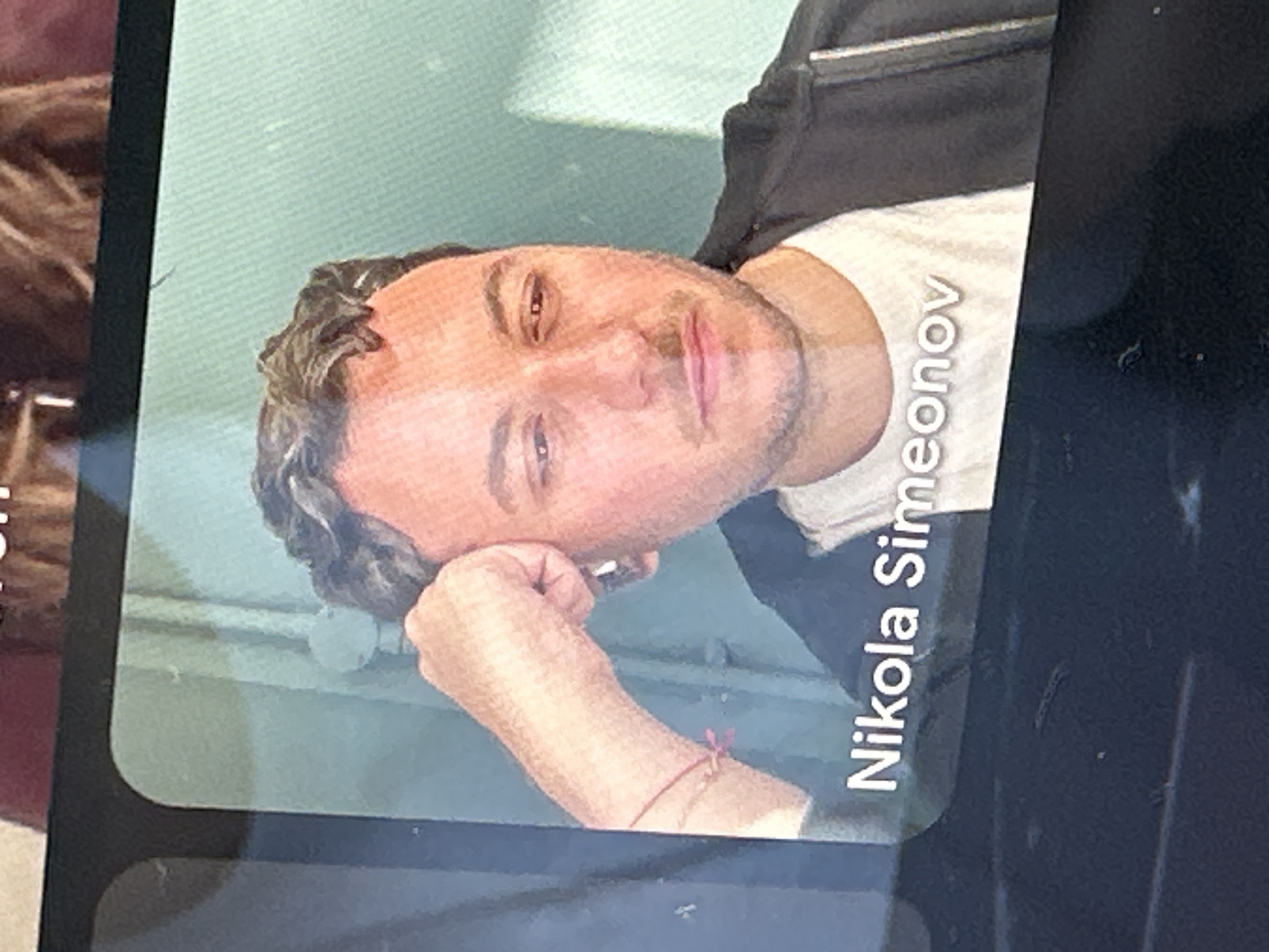 An image, captured from an angle, displays a man on a digital screen, likely a photograph of a screen rather than the original image itself. The man, identified by text as Nikola Simeonov, has a light complexion, dark wavy hair, and a light beard/stubble. He wears a white t-shirt and a dark jacket or hoodie. His left arm is bent, with his hand resting near his face in a relaxed, contemplative pose. A thin red string bracelet with a small knot is visible on his left wrist. He looks directly at the camera with a calm and composed expression.
The background visible on the screen behind the man is a plain, light blue-green wall or surface, suggesting an indoor setting. Faint horizontal lines can be seen in this background. The overall image captures the screen within a darker, out-of-focus environment; a blurred part of what appears to be another person's head with brown, curly hair is visible on the far left edge of the original photo.
The most prominent visible text is "Nikola Simeonov," written in white capital letters on the man's white t-shirt, oriented vertically along his right side (due to the image's rotation). No specific time of day or weather conditions are discernible. The image does not provide visual cues for the specified location of London, United Kingdom, which is provided as external context.
