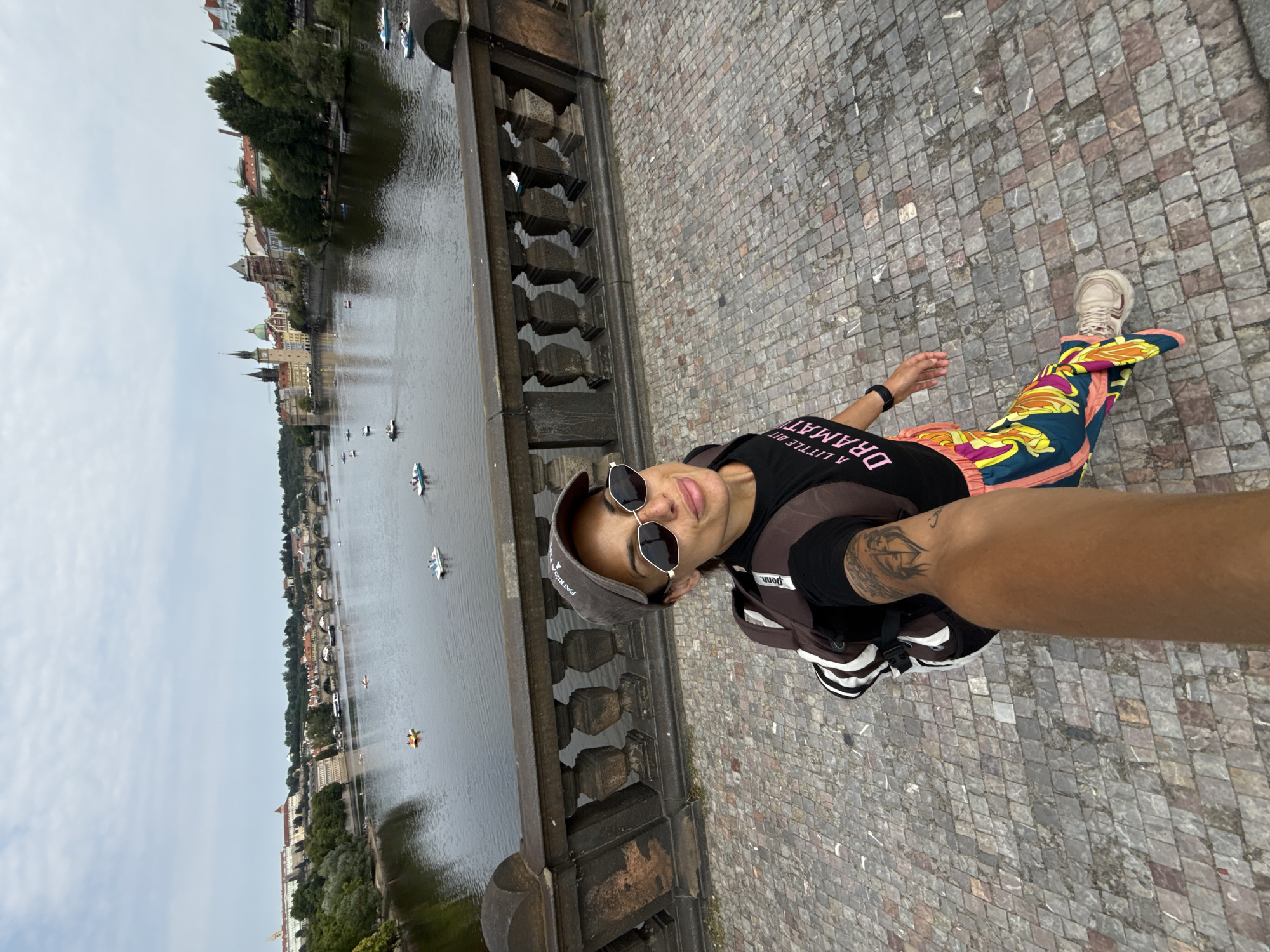 A woman wearing a black t-shirt with pink text, sunglasses, and a grey baseball cap takes a selfie while standing on a cobblestone bridge in Prague, Czechia. She is also wearing a backpack and brightly colored pants. In the background, the Vltava River is visible with several small boats on its surface and the cityscape of Prague with its historic architecture. The sky is overcast.