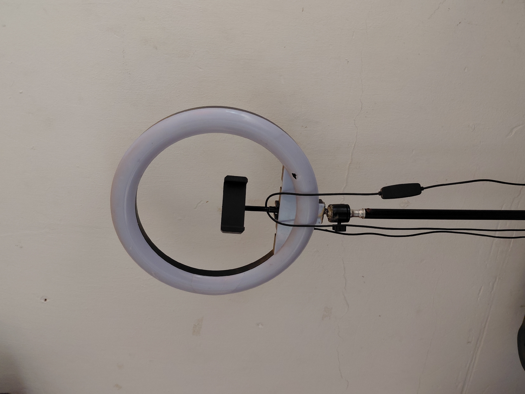 The image displays a white LED ring light, actively illuminated, positioned against a light grey, textured wall. A black smartphone holder is centrally mounted within the ring light's aperture. To the right of the ring light, a black pole or stand extends horizontally. Black cables are connected to the ring light and run along this pole, featuring a black, rectangular control switch on one of the cables. The wall, which acts as the background, shows some minor blemishes and subtle surface cracks. No individuals are present in the frame, and no specific activity is taking place beyond the ring light being switched on, suggesting a ready setup for photography or video recording. The time of day and external weather conditions are not discernible from the image. The setting appears to be an indoor space. There is no visible text.