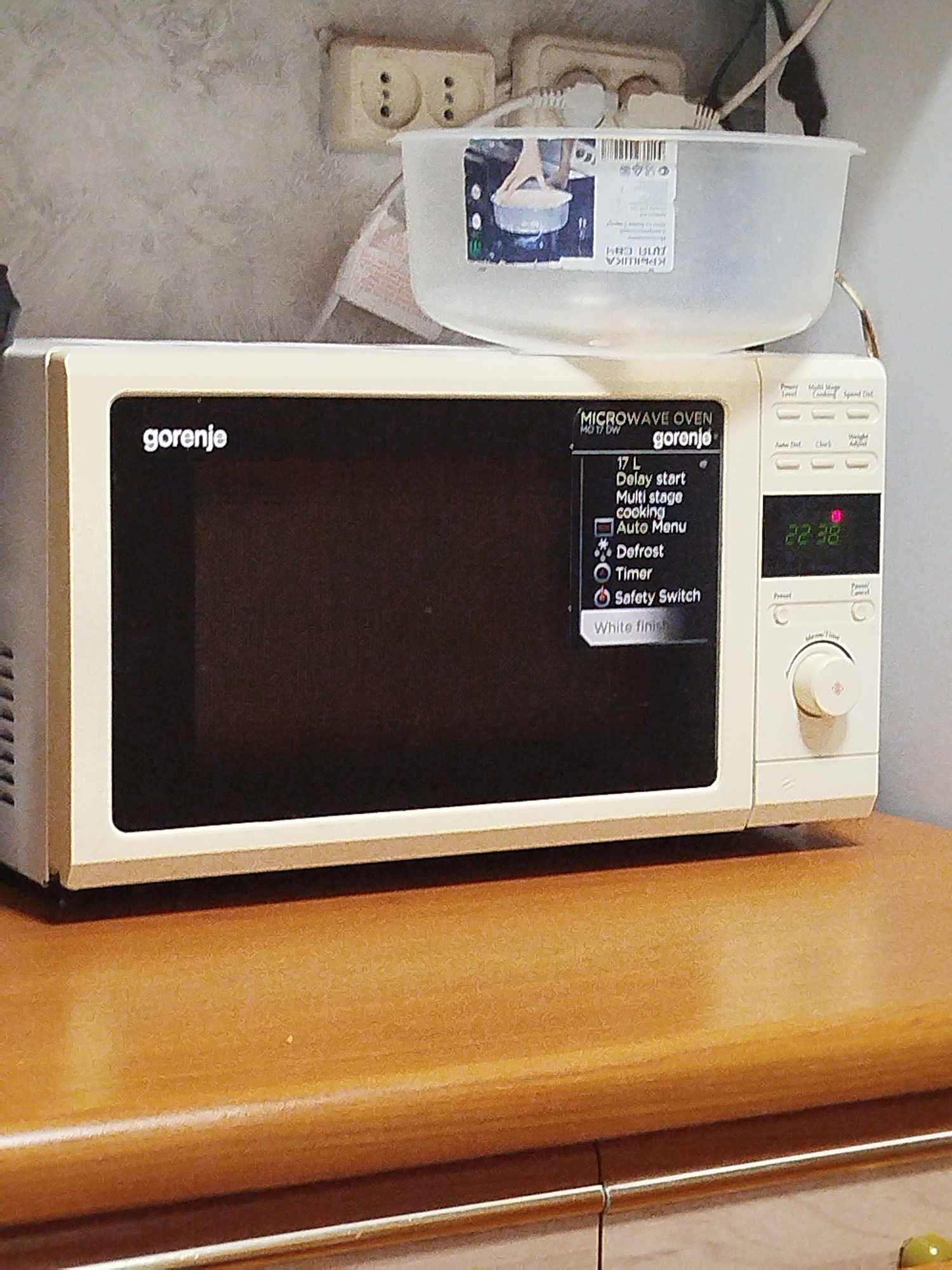 The image shows a Gorenje microwave oven, model MD 17 DW, with its control panel displaying "22:38". The microwave is positioned on a wooden surface, likely a countertop or shelf. Above the microwave, a white plastic container is visible, and behind it, an electrical outlet with two sockets is mounted on a textured wall. The overall environment appears to be indoors, possibly a kitchen. There are no people or actions depicted in the image. The city of Шой-Шудумарь, Russia is mentioned but not directly depicted or relevant to the visual content of the image.