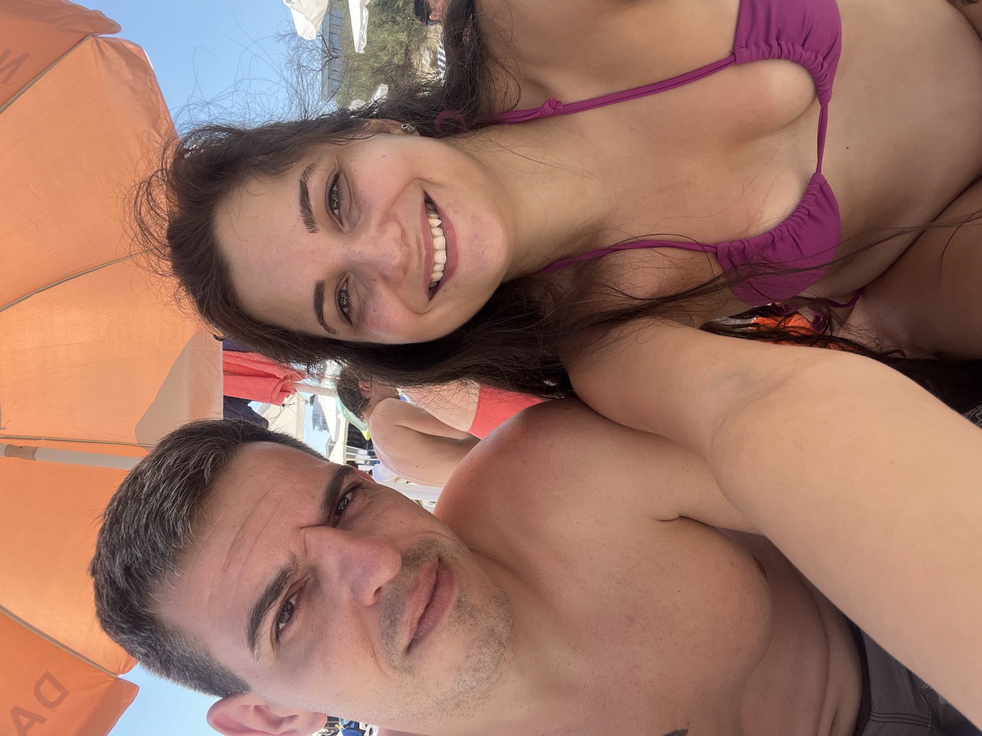In Pomorie, Bulgaria, a couple is taking a selfie on a beach, likely under an orange umbrella for shade. The man, on the left, is shirtless, with short, dark hair and a stern expression, looking directly at the camera. The woman, on the right, is wearing a purple bikini top and is smiling broadly, also looking at the camera. Her long, dark hair is slightly windswept. In the background, other beachgoers and white umbrellas are visible, suggesting a busy day at the seaside. The lighting indicates it is daytime, and the clear blue sky suggests fair weather.