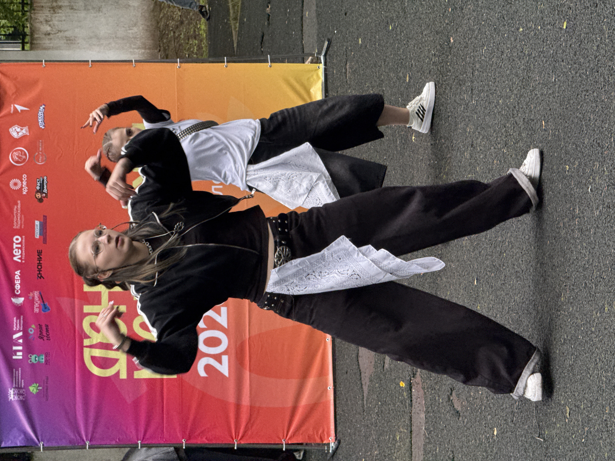 The image shows two young women dancing in front of a banner. The banner has "202" visible, suggesting a year. There are logos on the banner, including one that appears to say "ДМИТРОВ" (Dmitrov), indicating the location is Dmitrov, Russia. The woman in the foreground is wearing a black jacket, black pants, and white sneakers. She has glasses and her hair is in braids. The woman in the background is wearing a white t-shirt, dark pants, and white sneakers. Both women are in motion, with their arms raised and hands in dynamic poses. The ground is asphalt. The lighting suggests it is daytime, possibly overcast.
