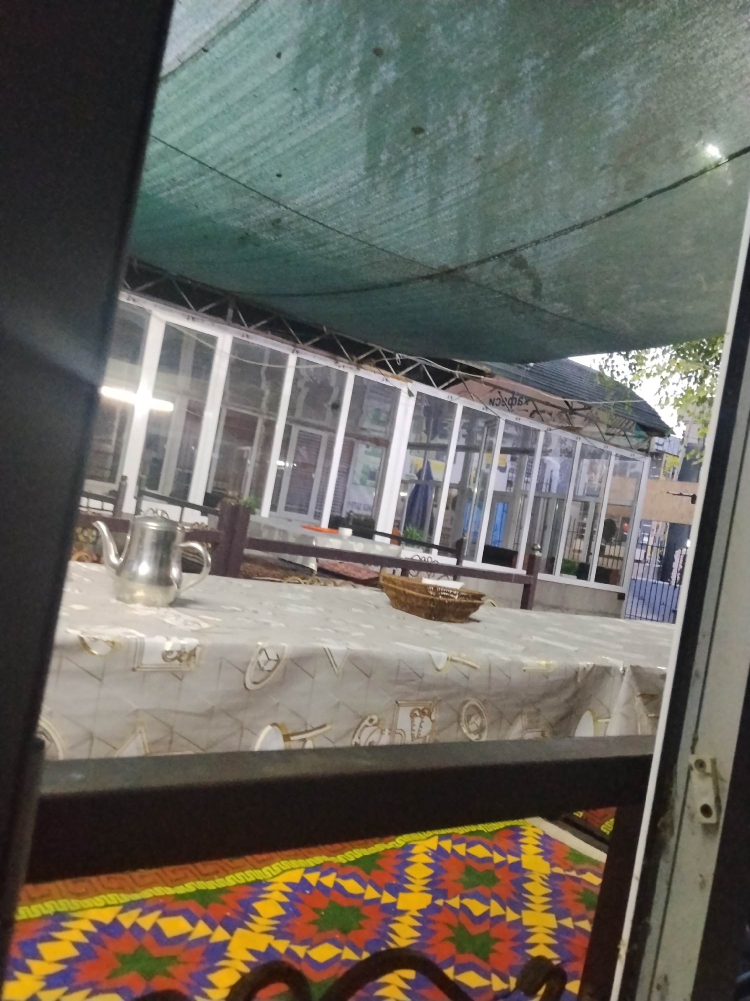 The image depicts an outdoor seating area of a cafe in Ala-Buka, Kyrgyzstan. A green mesh canopy is stretched overhead, providing shade. In the foreground, a table is set with a tablecloth featuring a pattern of golden decorative elements and clocks. On the table, a silver teapot and a woven basket are visible. Behind the table, a glass-enclosed building is seen, likely part of the cafe's interior, with multiple windows and doors. The overall impression is of a simple, functional outdoor dining space.