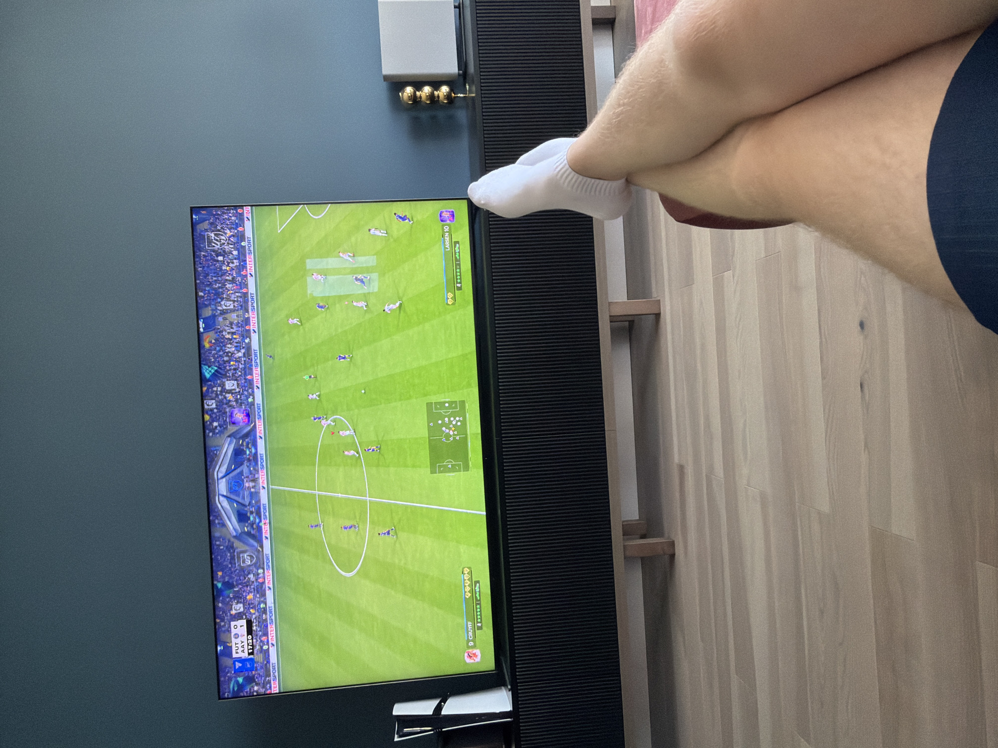 The image shows a person's legs, crossed at the ankles, with white socks and dark blue shorts visible. They are sitting in front of a large television screen displaying a soccer video game, likely FIFA, with an in-game score of 0-1 and the time at 17:20. The TV is on a black media console with a light wood base. To the left of the TV is a white gaming console. Behind the TV is a dark teal wall. To the right of the TV, on the console, is a decorative object with three gold spheres and a white rectangular object. The floor is light-colored wood. There is no indication of the location being Kyiv, Ukraine.