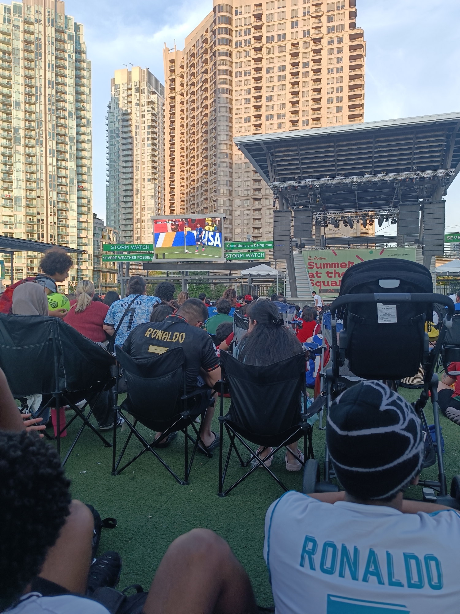 The image depicts a crowd gathered in an outdoor urban setting, likely at Celebration Square in Mississauga, Canada, to watch a soccer game on a large screen. Towering residential buildings frame the scene, characteristic of a downtown area. People are seated in portable chairs on a green, artificial turf surface. In the foreground, individuals are focused on the game being broadcast. Some are wearing jerseys with the name "RONALDO" prominently displayed. A stroller is visible, suggesting families are present. The overall atmosphere appears to be one of communal enjoyment of a sporting event in a public urban space. A banner reading "Tim Hortons Summer at the Square" is visible, indicating a community event. The sky is partly cloudy.