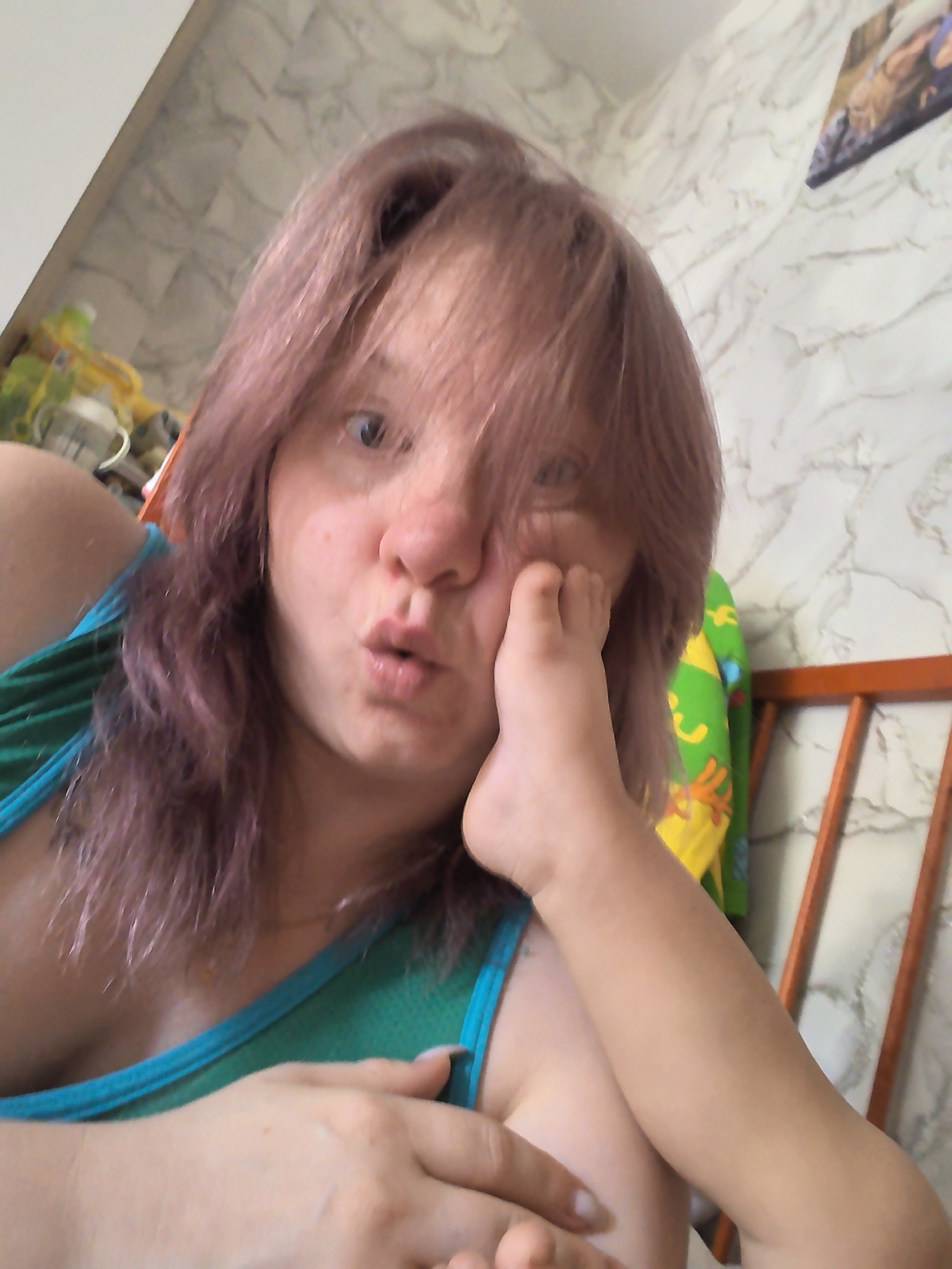 A close-up, overhead shot captures a person with pinkish-purple hair and fair skin looking directly at the camera with a surprised expression, their mouth slightly open. The person is wearing a teal tank top. A child's foot is positioned on the person's cheek, obscuring part of their face. In the background, a wall with a marble-like pattern is visible, along with a section of a wooden chair and a colorful, cartoon-patterned fabric. There is no specific indication of the city of Chaikovsky, Russia, in the image.