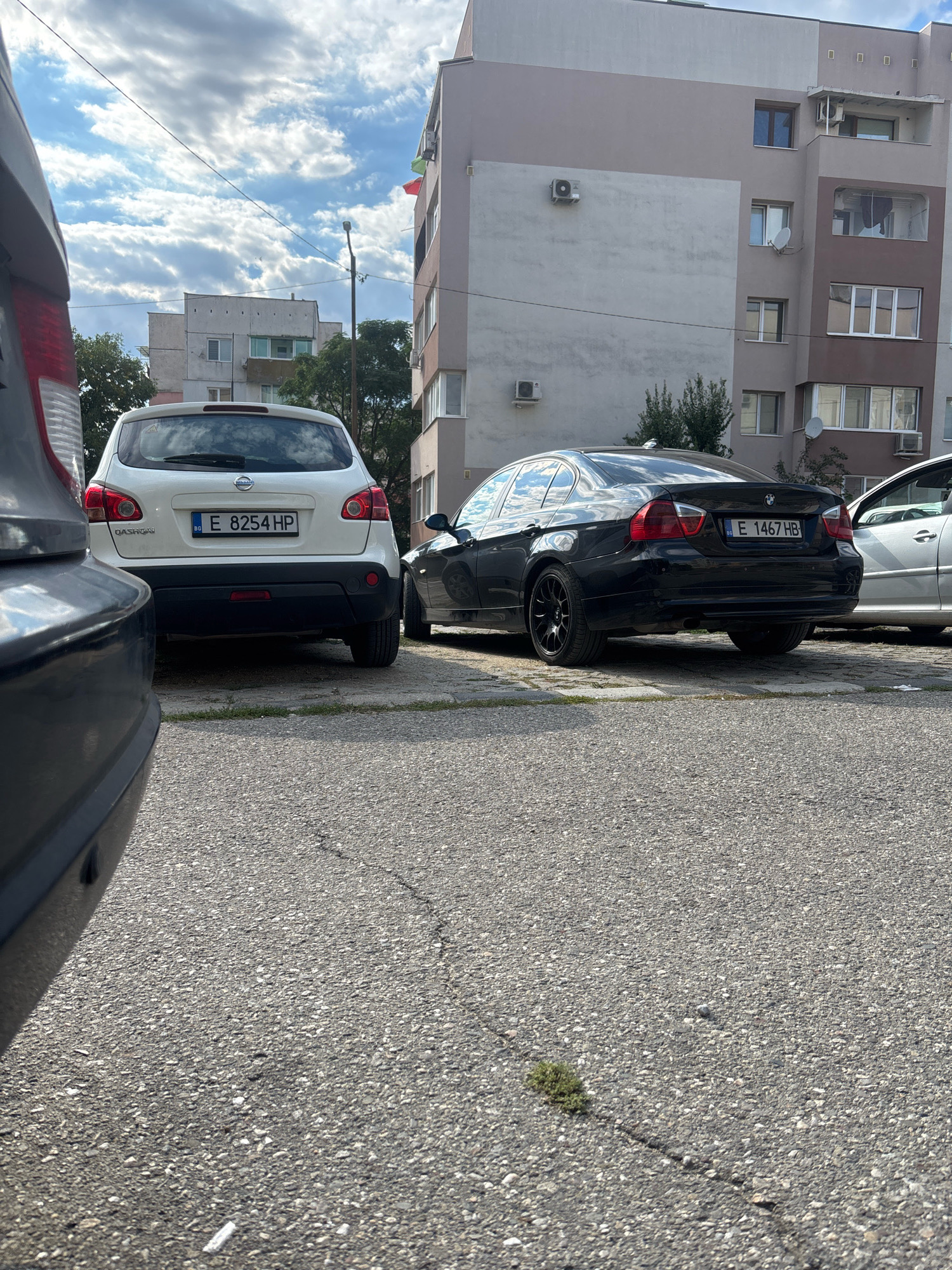 The image shows a street scene in Blagoevgrad, Bulgaria. In the foreground, a dark-colored car is parked on asphalt pavement. To the left, a white Nissan Qashqai is parked, with its rear visible, displaying the license plate "BG E 8254 HP". To the right, a black BMW sedan is parked, with its rear visible, displaying the license plate "BG E 1467 HB". Further to the right, a silver car is partially visible. Behind the cars, a multi-story apartment building dominates the background, with several windows and balconies. The sky is partly cloudy, with a lamp post visible between the cars and the building. The overall impression is a typical urban streetscape.