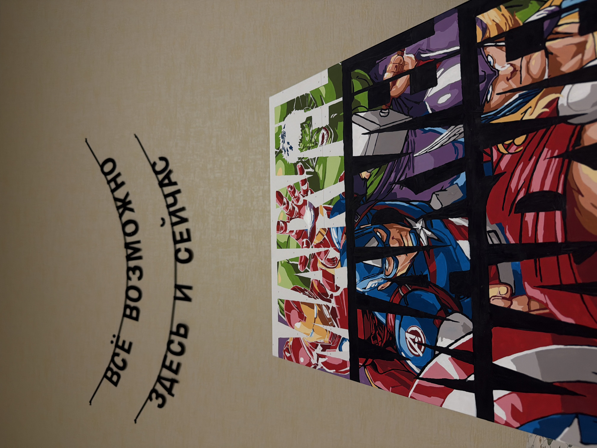 The image shows a mural on a wall. On the left side of the image, there are black Cyrillic letters arranged in two lines. The first line is curved upwards. A black line intersects both lines. The phrase translates to "Everything is possible here and now." To the right is an Avengers themed artwork, divided into strips by thick black lines. The visible characters include Hulk, Iron Man, Captain America, and Thor. The mural is presumably located in Izhevsk, Russia.