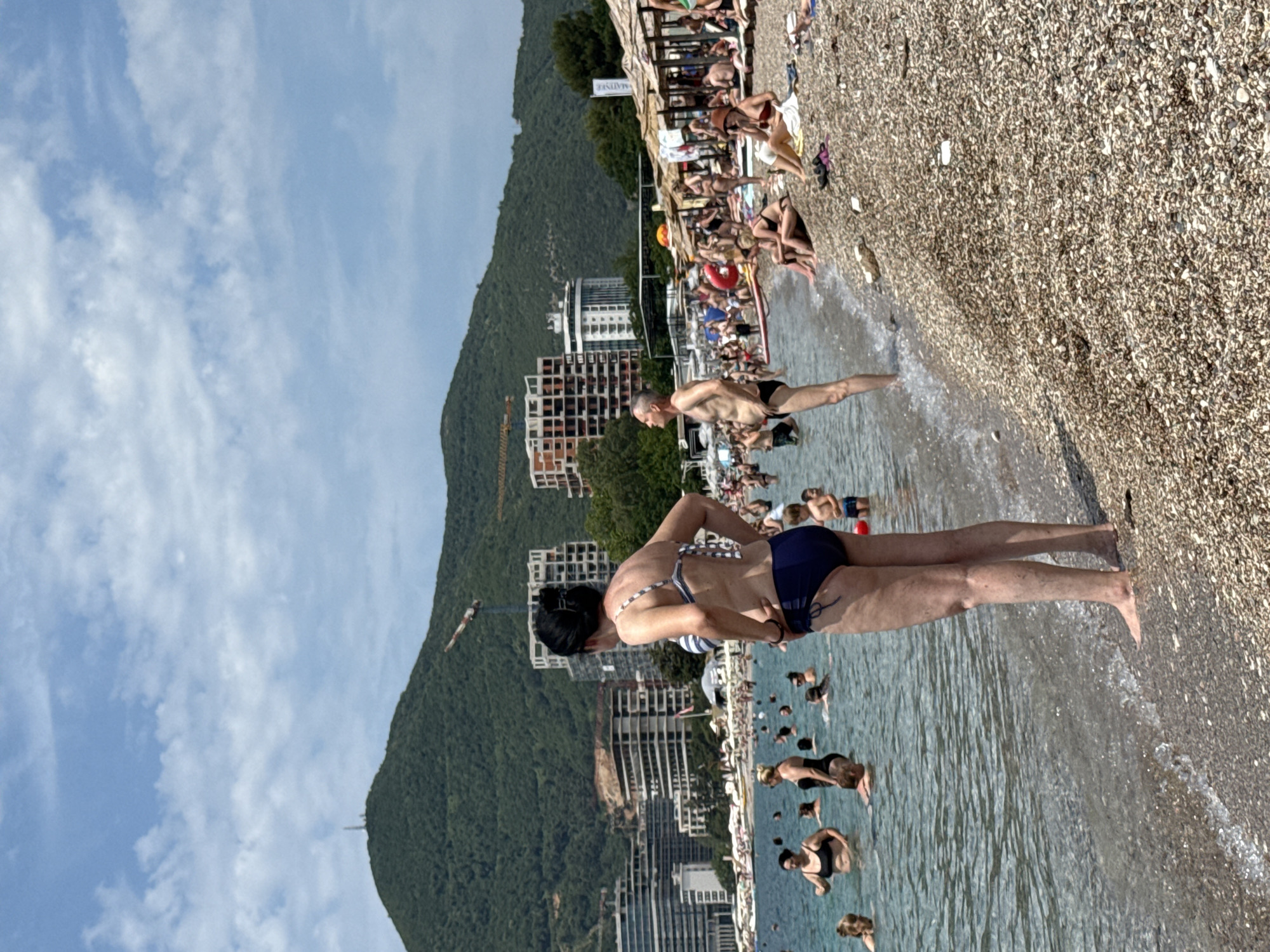 The image depicts a crowded beach in Boreti, Montenegro, on a sunny day with a clear blue sky and scattered clouds. The foreground shows a woman in a blue and white striped bikini standing in the shallow water, facing away from the camera. The water is clear with small waves lapping at the pebble beach. Behind her, other beachgoers are visible, some in the water and some on the shore. A man in black swimming trunks stands in the water to the right of the woman. Further back, a stretch of beach lined with thatched umbrellas is occupied by many people sunbathing and relaxing. In the background, the coastline is dominated by lush green mountains, with modern buildings and construction cranes indicative of urban development. The sea stretches out to the horizon.