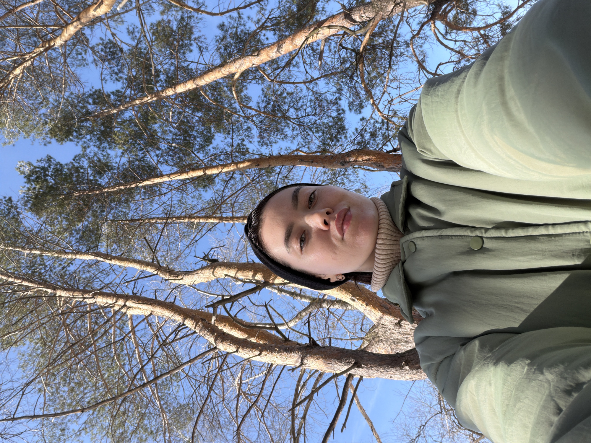 A low-angle selfie depicts a young adult female positioned outdoors in the city of Chaikovsky, Russia. The subject occupies the lower half of the frame, facing the camera directly with a neutral expression and slightly pursed lips. She wears a black headband, a ribbed beige turtleneck, and a loose-fitting olive-green collared jacket with light-colored buttons. A small, light-colored earring is visible on her right earlobe. The background consists of several tall pine trees with textured light brown bark and numerous branches, some appearing bare and others retaining sparse green needles. These trees ascend towards a clear, bright blue sky. The scene is illuminated by natural sunlight, indicating daytime.