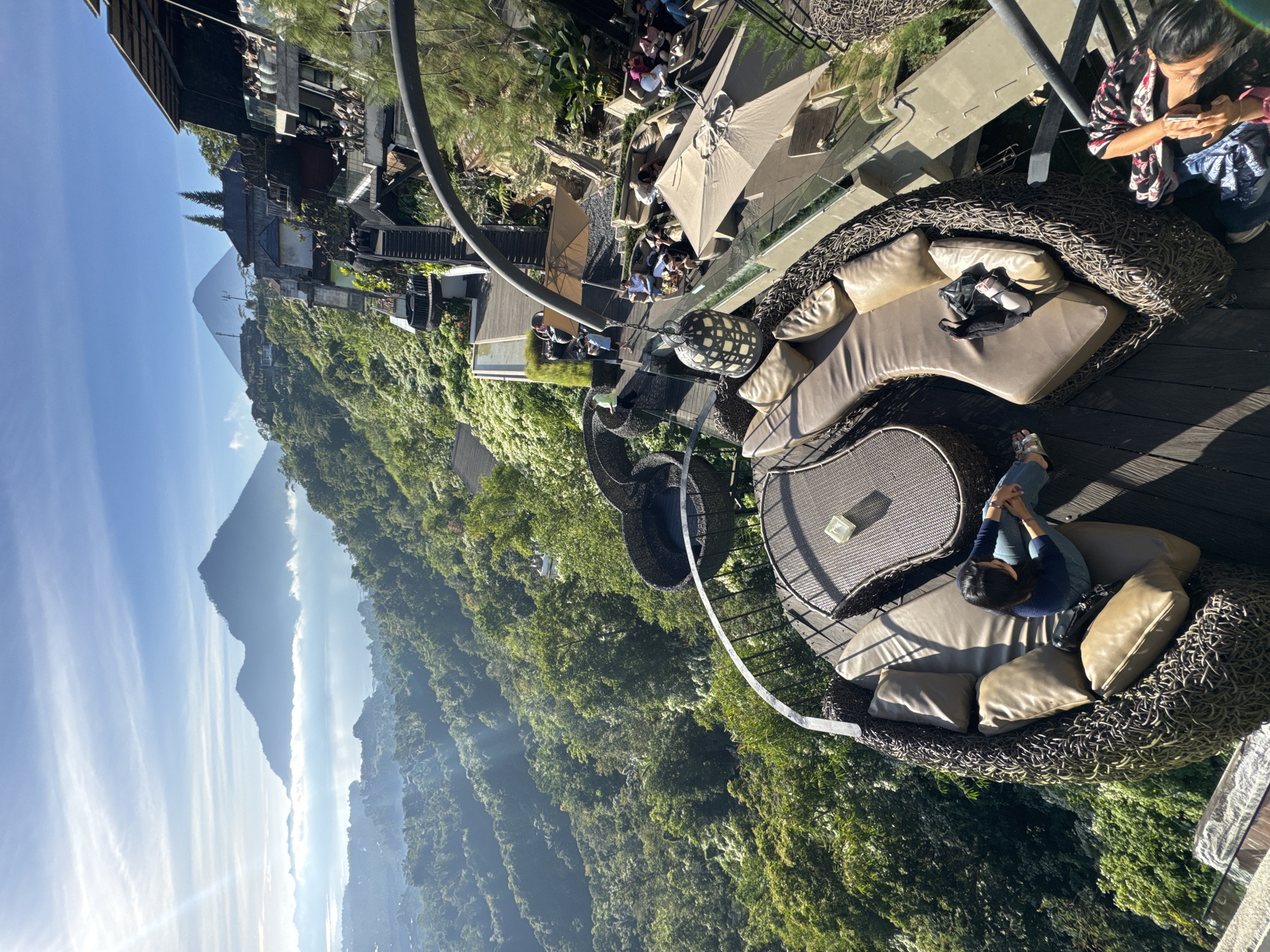 A high-angle, long shot captures a panoramic view of a mountainous landscape in Kintamani, Indonesia, bathed in bright, morning sunlight. In the foreground, a person is seated on a large, circular rattan sofa with plush cushions, overlooking the expansive vista. The sofa is part of an outdoor seating area on a wooden deck, adorned with a decorative lantern. Beyond the deck, a multi-level establishment built into the hillside features various seating areas with umbrellas and railings, occupied by diners. A winding staircase connects the different levels. The background showcases a vast expanse of lush, green forest cascading down steep slopes, leading to a misty lake and two prominent mountains in the distance, partially obscured by clouds. The sky is a clear blue, with streaks of white clouds and lens flare from the sun.