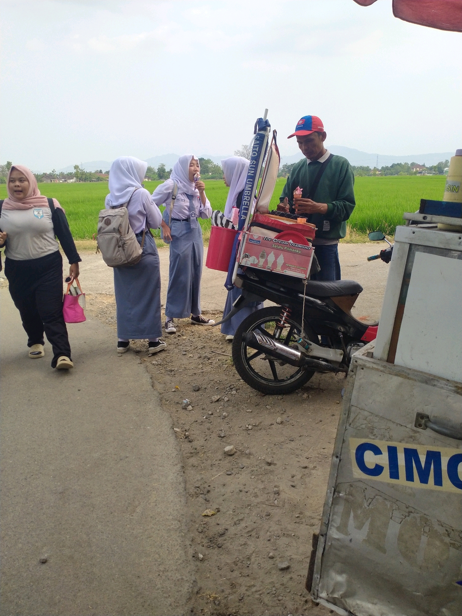 This is a waist-up, eye-level, medium shot of an ice cream vendor and several students in Begajah, Indonesia. The vendor, a man in a green long-sleeved shirt and a red and blue baseball cap, stands to the right, holding an ice cream cone. He is next to a motorcycle with a sidecar ice cream cart. The cart has a box of ice cream with images of ice cream cones and text that says "Ice Cream Diamond Selalu Pilihanku". To the left of the vendor, three young women in school uniforms and headscarves are visible. One woman in the foreground, wearing a light pink headscarf, is walking away from the viewer. Two other women, in white headscarves and blue school uniforms, are standing closer to the cart, one of whom is eating an ice cream cone. The background features a lush green rice paddy under a bright, overcast sky. The scene is set on a dirt and asphalt road.