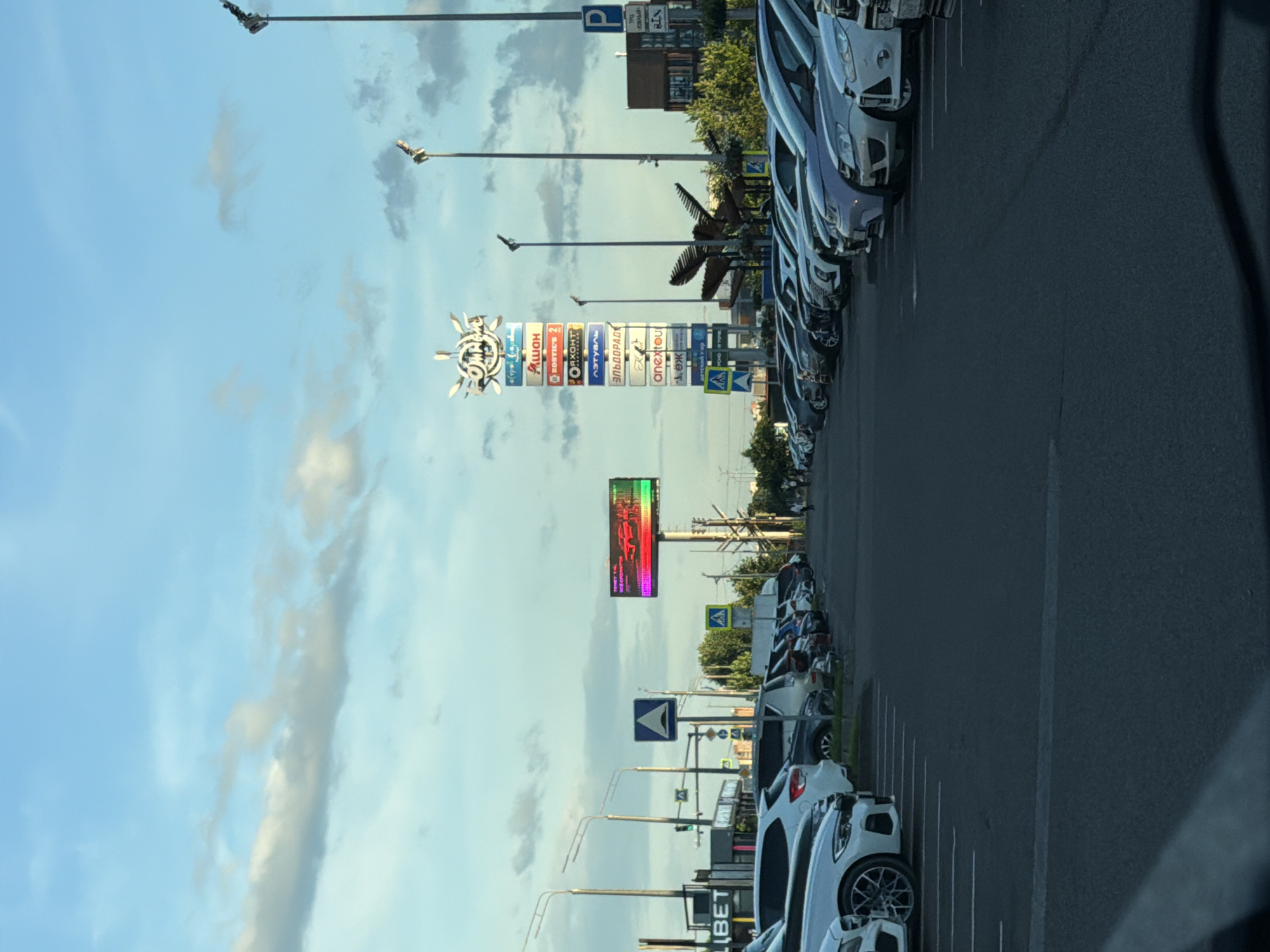 This is a vertical, medium shot photograph of a parking lot filled with cars. The sky is blue with some scattered clouds, suggesting it is daytime. The lighting indicates it might be late afternoon or early evening.
In the foreground, a row of cars is parked on asphalt. The cars are of various colors, with several white and silver vehicles prominently visible. The parking lot is marked with white lines.
In the background, there are several tall lampposts, typical of a parking lot or outdoor area. There is also a tall signpost with multiple rectangular signs advertising various businesses. Some of the text on these signs is in Cyrillic script, and includes words like "Ашан" (Auchan), " Эльдорадо" (Eldorado), and "oneTwo" (oneTwo). A large, colorful digital billboard is also visible, displaying a dynamic image. Further back, more trees and buildings can be seen. The overall scene depicts a commercial area, likely a shopping center or retail park. No people are clearly visible in the image.