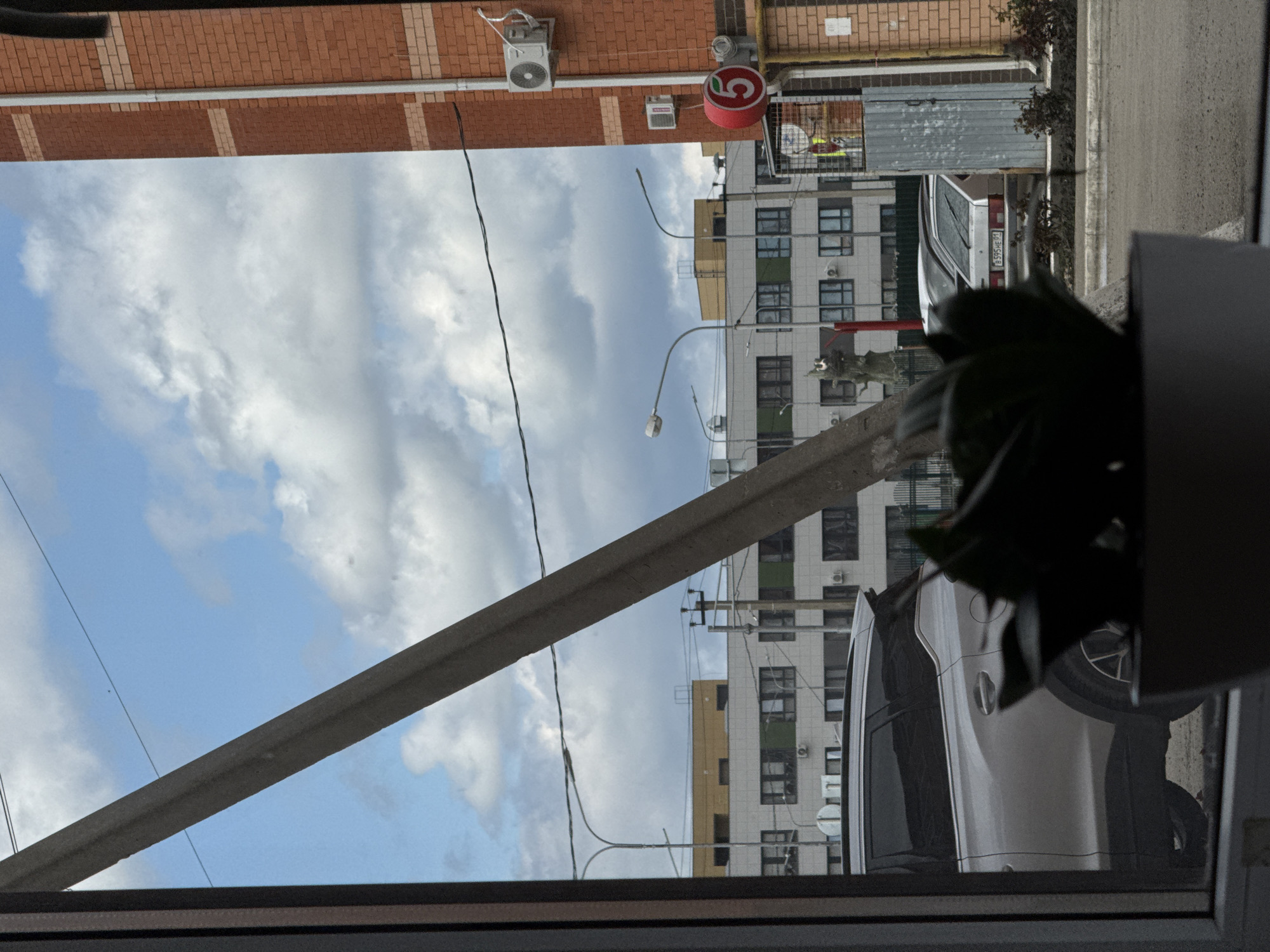 The image shows a view from a window, likely in an urban setting in the city of Unknown, Unknown. The lower part of the frame shows the window frame and part of a plant. Outside the window, there is a white car parked next to a building with multiple windows. The building is mostly white with some yellow sections and multiple air conditioning units on the exterior. A street sign with the number "5" in red is visible. A brick building is also visible in the background. Power lines cut across the sky, which is partially cloudy. A concrete beam extends from the bottom left to the middle of the frame.