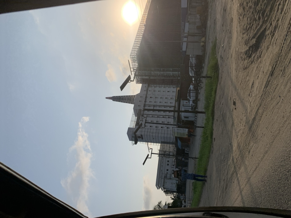The image depicts an urban or semi-urban street scene in Ikota, Nigeria, under a pale blue sky with scattered clouds and a bright, low sun visible on the far right, suggesting late afternoon or early evening. The foreground features an unpaved, sandy-dirt road exhibiting tire tracks and a strip of green grass on the left.
In the mid-ground, a lone male figure walks leftward across the dirt road, carrying a dark, round object balanced on his head. He wears a plaid shirt and dark trousers. Further back and to his right, another less distinct individual is visible near a white, shed-like structure.
The background is dominated by several multi-story buildings. To the left-center, a light-colored building, possibly white or light grey, is partially covered in scaffolding, indicating construction or renovation. A distinctive, dark, spire-like structure, reminiscent of an oil derrick, rises from its roof. To the immediate right of this building, a large, dark-colored structure is heavily covered with dark netting and scaffolding, signifying extensive construction. A sign with "BEAUTY CENTER" is partially visible on this structure's lower facade. Several white sedans and other vehicles are parked in an unpaved lot in front of this large dark structure.
Multiple tall poles with solar panel fixtures are positioned throughout the scene, presumably for lighting. Patches of sparse green vegetation are present near the buildings and along the road. The extreme upper left and right corners of the image show parts of a dark, curved frame, likely from inside a vehicle, indicating the photograph was taken from a moving or parked car.