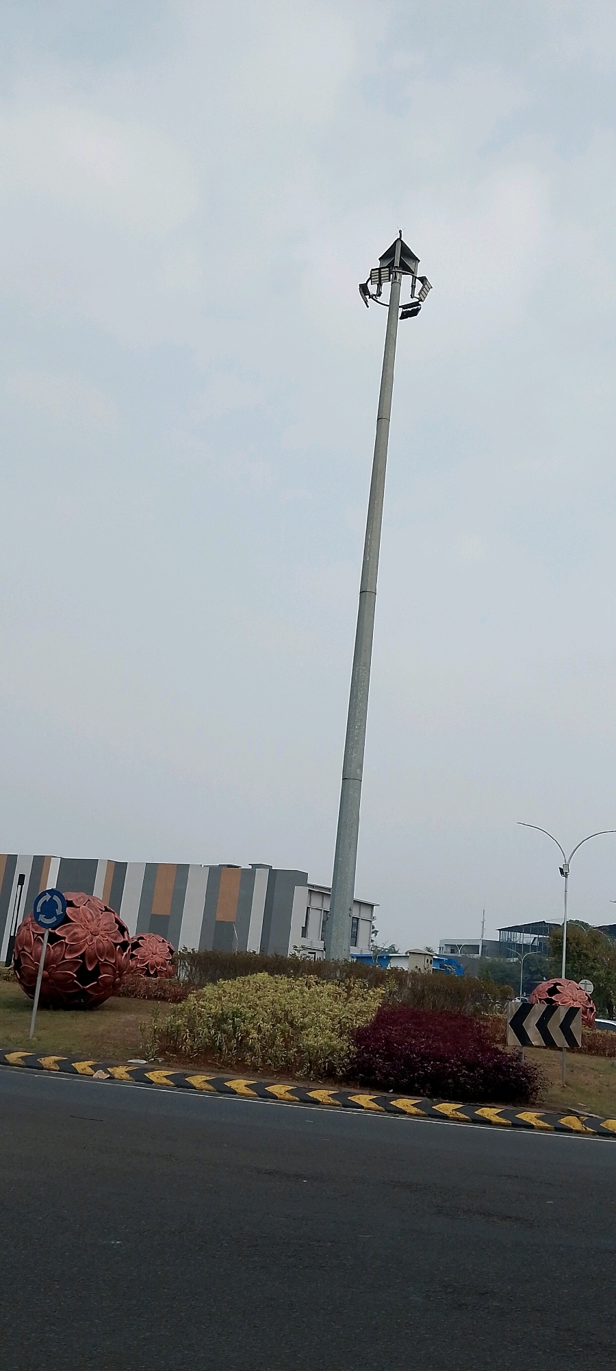 In Gunung Putri, Indonesia, a tall, slender pole with multiple lights stands at the center of a roundabout. The roundabout itself is adorned with large, decorative spherical objects that appear to be made of metal and are intricately designed with petal-like patterns. These spheres are positioned around a circle of low-lying green and red shrubs, bordered by a road with distinctive yellow and black striped markings. In the background, a modern building with a facade of vertical grey, white, and orange stripes can be seen. The sky above is overcast with a pale, hazy cloud cover.