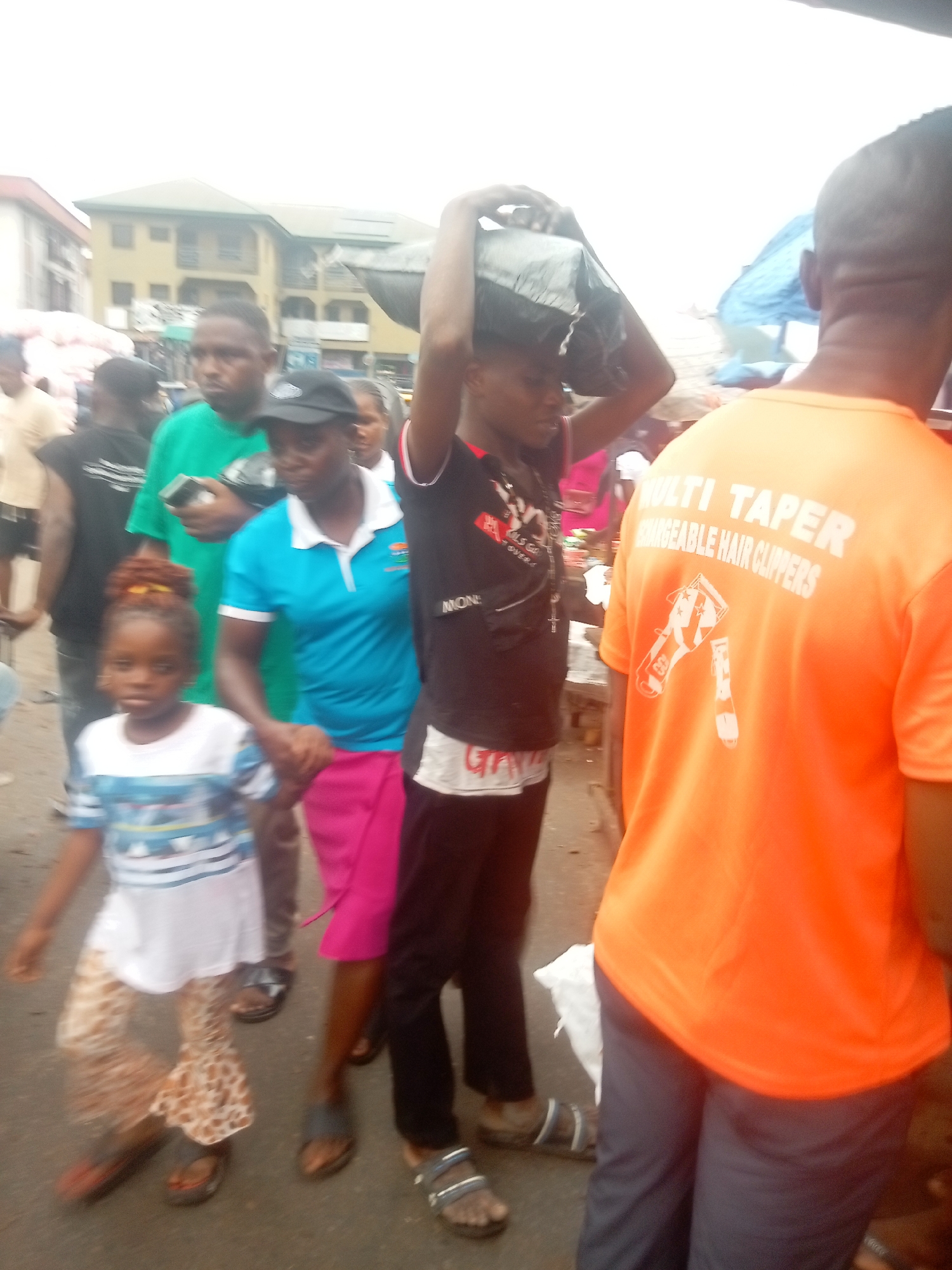 The image captures a street scene in Onitsha, Nigeria, evidenced by the architecture and the clothing styles of the people. A diverse group of individuals are visible. In the foreground, a child in a patterned outfit walks alongside a woman in a blue shirt and pink skirt. Beside them, a man carries a dark bag atop his head. To the right, another man wears an orange shirt printed with "MULTI TAPER RECHARGEABLE HAIR CLIPPERS". Several other individuals populate the background, including a man in green using a mobile phone. The setting suggests a bustling marketplace or public area.