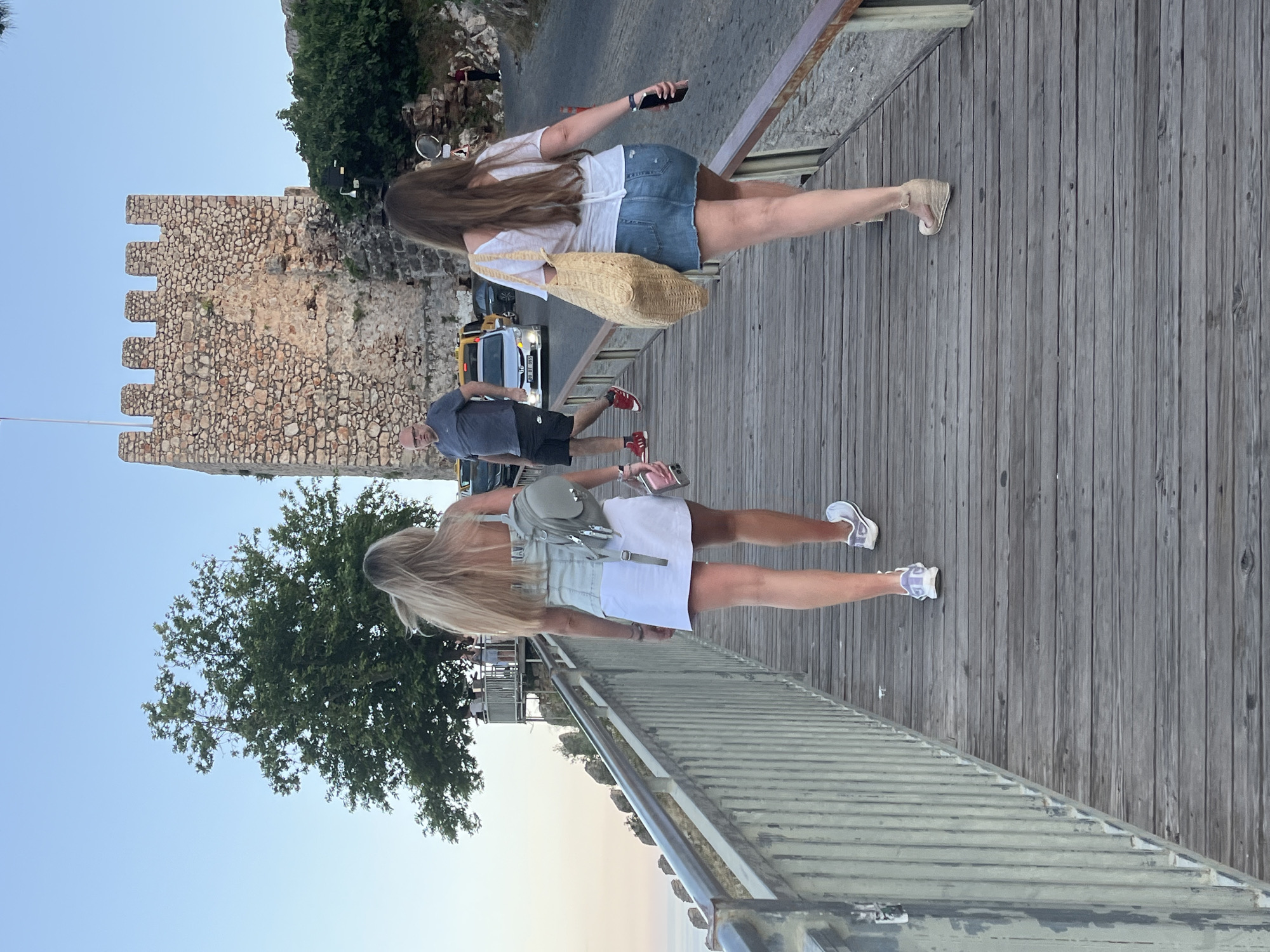 A vertically oriented, eye-level photograph captures a scene from Alanya, Turkey. Two women and a man are walking on a wooden walkway. The woman in the foreground has blonde hair and wears a light-colored dress with a backpack. The woman behind her has long brown hair, wearing a white top and denim shorts, carrying a woven bag, and holding a smartphone. A man walks between them. A stone structure with crenellations is visible behind the walkers, along with trees, a street, and vehicles. The background shows a gradient of blue and orange, indicating twilight or sunset. The scene suggests a public walkway, possibly a promenade or a bridge.