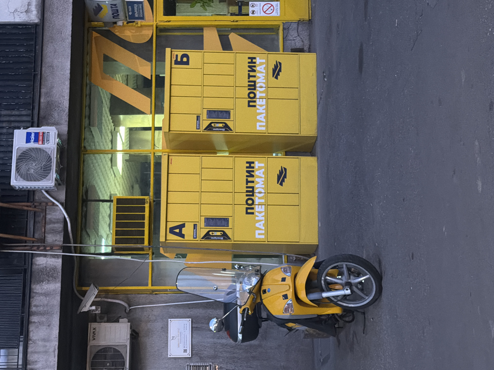 The image shows a street scene in Belgrade, Serbia, with a focus on a yellow "ПОШТИН ПАКЕТОМАТ" parcel locker. The locker is labeled with the Cyrillic letters "A" and "Б," corresponding to English "A" and "B." The locker is located next to a building with a Haier air conditioner unit and a security camera. There is also a yellow moped parked in front of the locker. Above the parcel locker, there is a sign with "ПОШТА" written on it.