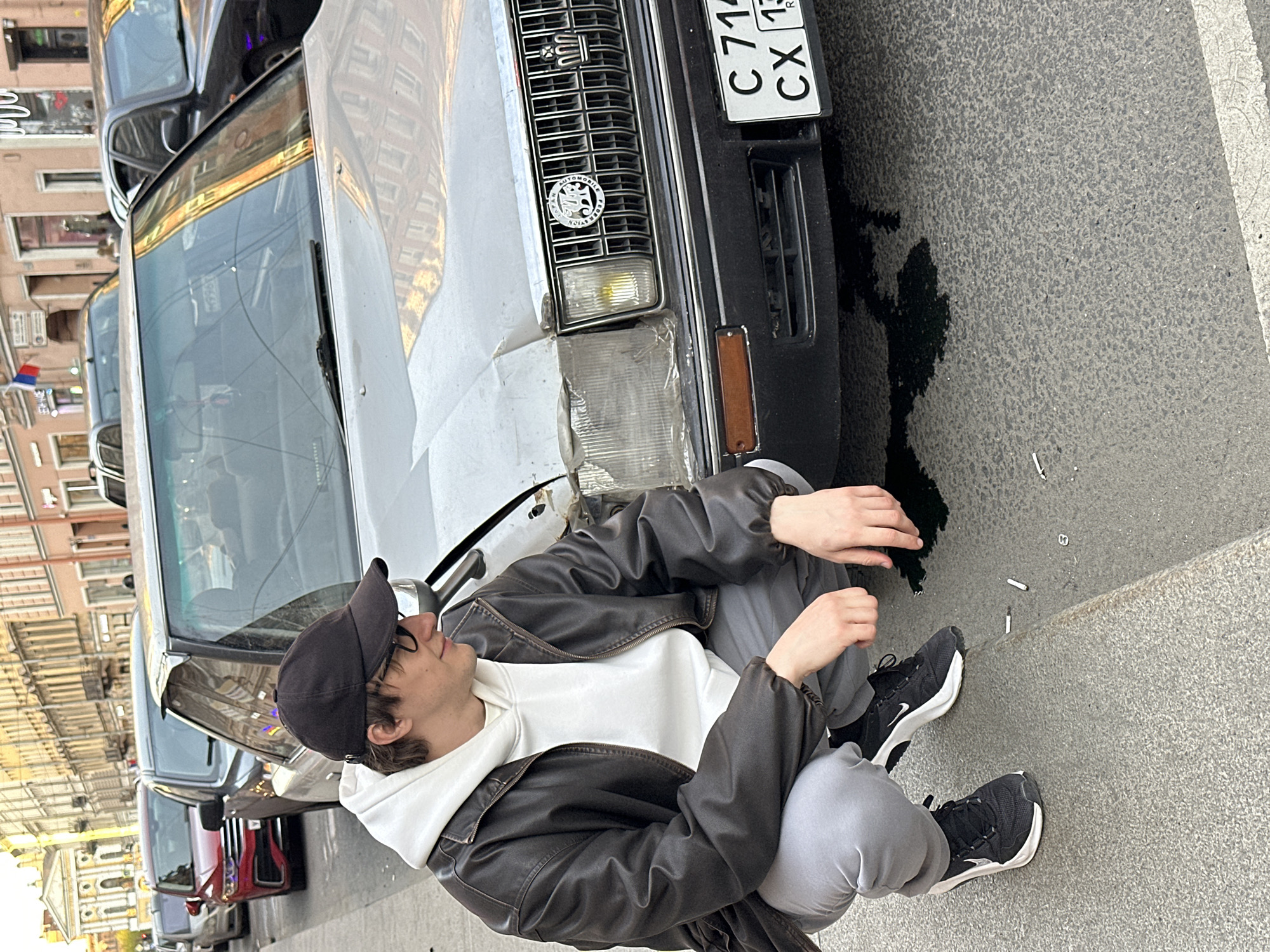 A young man is captured squatting on a paved street next to the front of a light-colored, older model car in Saint Petersburg, Russia. He is wearing a black baseball cap, glasses, a dark brown leather jacket over a white hoodie, grey sweatpants, and black sneakers. His gaze is directed downwards, possibly at the car's front or the ground.
The car's front bumper and driver's side headlight area show visible damage, with the headlight assembly appearing taped or covered. A dark fluid, likely oil or coolant, has leaked onto the asphalt directly beneath the car's front. The vehicle features a distinctive chrome grille and an emblem that reads "AUTOMOBILE FEDERATION SYSTEM" and "AMERICAN HERITAGE". A Russian license plate, "C 714 CX 178 R", is attached to the front, with "178" being the region code for Saint Petersburg.
In the background, other vehicles are parked, and reflections of urban buildings with windows are visible in the car's windshield. Several cigarette butts are scattered on the ground near the man's feet. The scene depicts a daytime setting with clear weather on an urban street.