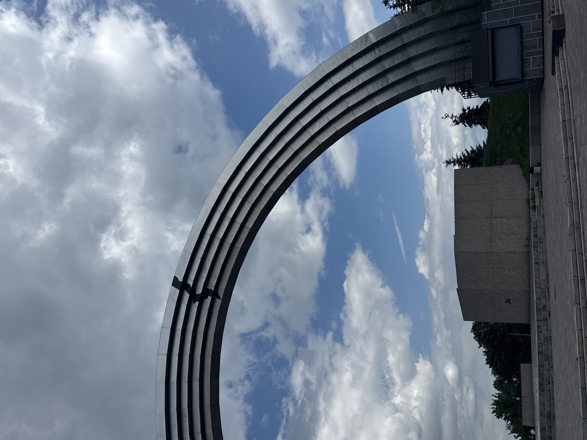 The image shows the Arch of Freedom in Kyiv, Ukraine, under a cloudy sky with patches of blue. The monumental arch, constructed from several curved metal sections, dominates the upper portion of the frame. A dark, jagged line, possibly a crack or a shadow, breaks across the topmost curved section of the arch. Below the arch, a grey, rectangular structure with a rough surface is visible, along with trees and a low, dark-colored kiosk. Paved pathways and steps lead towards these structures. In the distance, more trees and a slightly sloped grassy area can be seen. The overall scene suggests an outdoor public space, possibly a park or monument area, in a city.