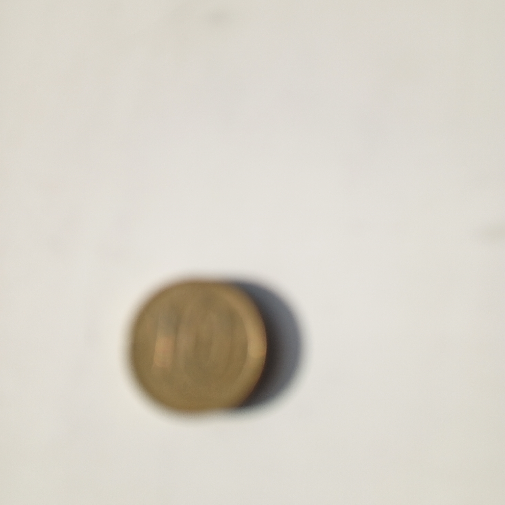 The image shows a single, out-of-focus coin lying on a light-colored surface, casting a subtle shadow. The coin appears to be gold or brass in color and has the numeral "10" visible on its surface. The background is a plain, textured off-white. There is no indication of the city of Sayanogorsk, Russia, or any other specific location. The image is a close-up of the coin and its immediate surroundings.