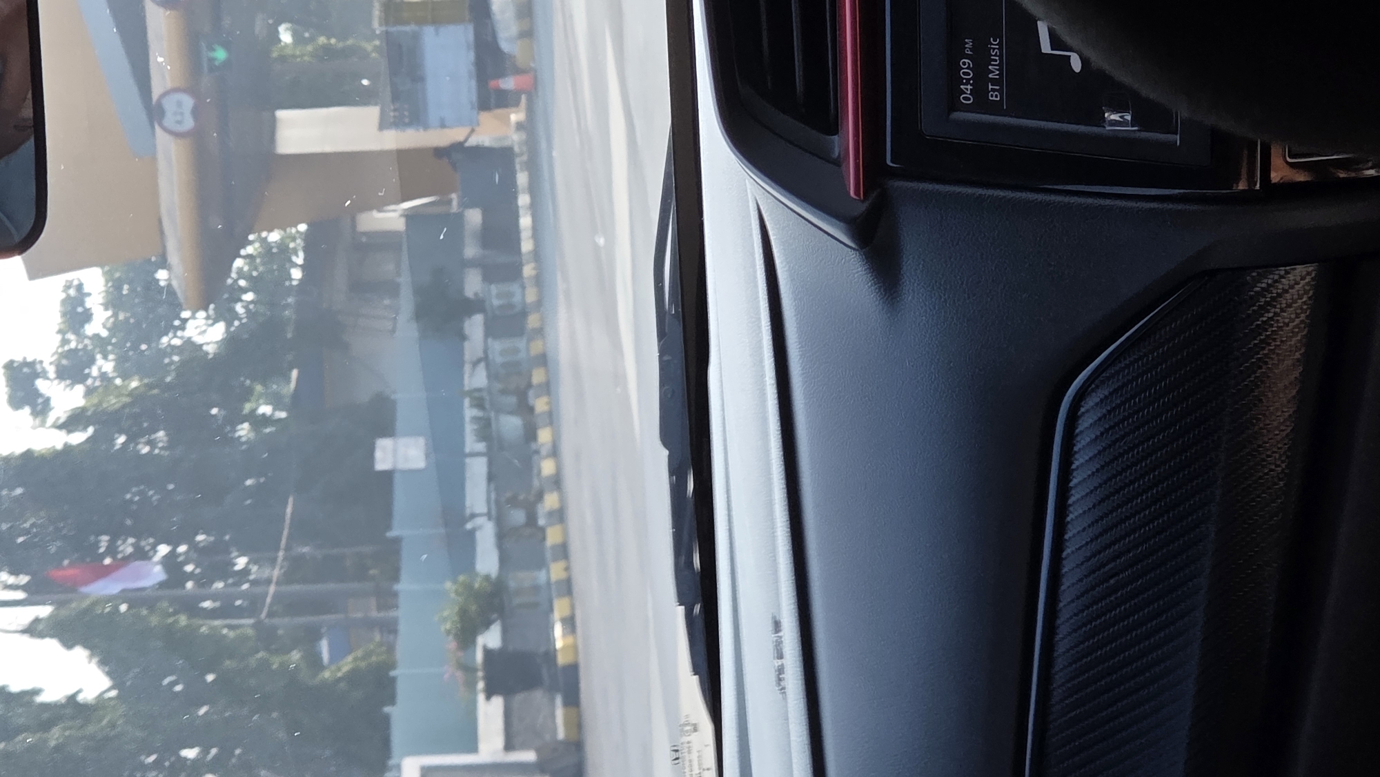 The image shows a view from inside a car, looking out through the windshield. The car appears to be approaching a toll booth. In the distance, an Indonesian flag flies on a flagpole. The car's dashboard is visible in the foreground, with a screen displaying "04:09 PM" and "BT Music." The street outside is lined with yellow and black striped barriers, and there are trees and buildings in the background. A toll plaza canopy with a height restriction sign of "4.2m" is visible, along with a green light indicating a lane is open. A traffic cone is present on the right side of the road. The scene takes place in Margajaya, Indonesia, during daylight.