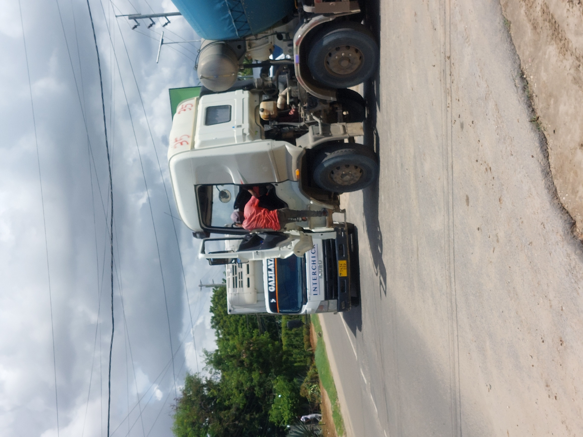 This image captures a street scene in Dar es-Salaam, Tanzania, featuring two trucks on a paved road under a cloudy sky.
In the foreground, a large white cement mixer truck is visible. A person wearing a red shirt is seated in the driver's cab. The side of the cab has the numbers "35 35" prominently displayed in red. The truck features a large blue mixing drum.
Behind the cement mixer, a white box truck or refrigerated truck is visible. This truck's cab features the text "INTERCHICK" and "CALLATAV" (partially visible) on its front grille. A yellow Tanzanian license plate, "T 434 DEP", is clearly affixed to the front bumper.
The road appears to be asphalt or paved, with a dirt shoulder on the right side. Beyond the road on the left, lush green trees and foliage are present. Utility poles with multiple power lines stretch across the frame against a heavily overcast, cloudy sky. Further down the road, near the trees, a faint figure of another person in light clothing can be seen standing by the roadside.
The scene appears to be taking place during the daytime, likely under overcast weather conditions, with no direct sunlight visible. The license plate and general setting are consistent with the provided location context of Dar es-Salaam, Tanzania.
