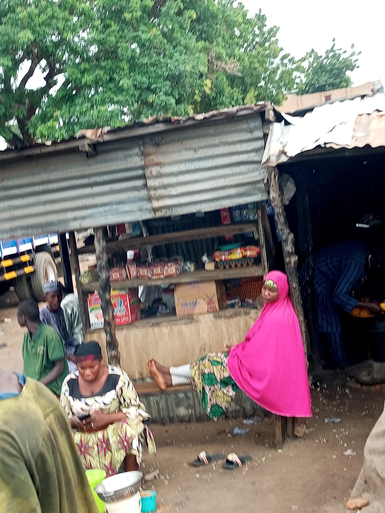 In Gadao, Nigeria, a street market scene unfolds under a corrugated metal roof supported by wooden poles. Several individuals are present. A woman wearing a bright pink hijab and a patterned dress is seated on a bench, leaning against a wooden post. To her left, a woman in a black, white, and yellow patterned dress is seated, looking at her phone. In the background, two men in green and grey shirts are standing, with a truck visible behind them. A shelf behind the seated women displays various items, possibly food products in colorful packaging. The ground is dirt, with some scattered debris.