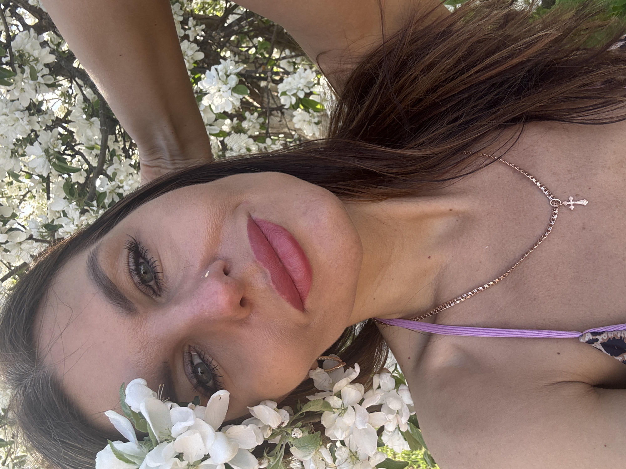A young woman with brown hair and fair skin is lying down in a field of white flowers. She is wearing a delicate gold necklace with a small cross pendant and a light purple strap of clothing visible on her shoulder. Her head is surrounded by the flowers, some resting on her cheek. The background is blurred, but suggests a natural setting with more white flowers and possibly trees. The photograph appears to be taken outdoors, possibly in a park or garden. The location is Dubovaya, Russia.