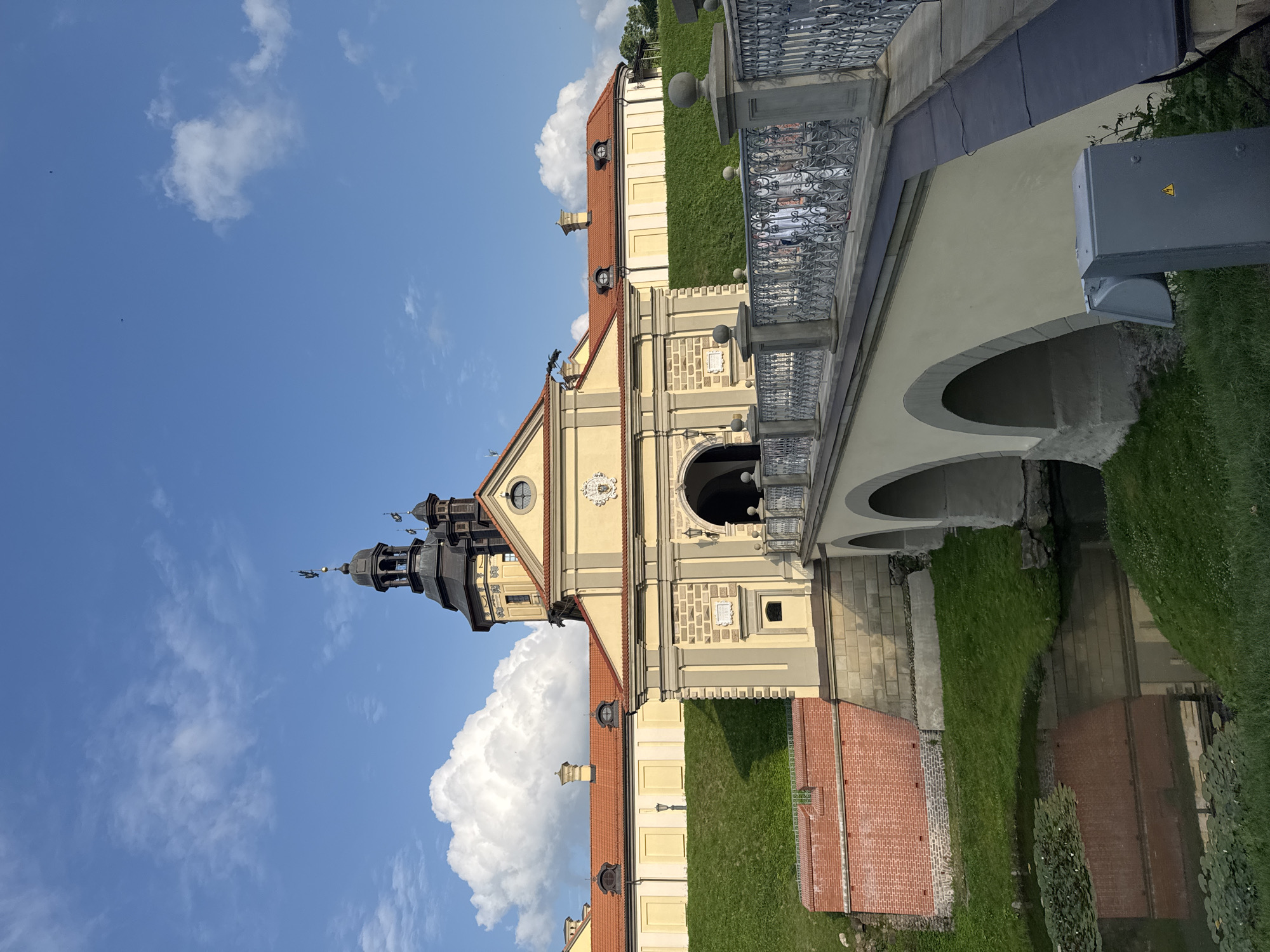 The image shows a sunny day at Nesvizh Castle, located in Nesvizh, Belarus. In the foreground, a stone bridge with arched supports spans a moat filled with lily pads. The bridge has an ornate metal railing with decorative finials. The bridge leads to a large, yellow, fortified building with a red tile roof. The building has a central archway and is adorned with decorative stone carvings. Behind the main building, several towers rise, including a tall, dark-colored tower with a spire and a weather vane. The sky is a vibrant blue with scattered white, fluffy clouds. Green grass covers the sloping hills surrounding the castle. A red brick wall is visible to the left of the bridge.