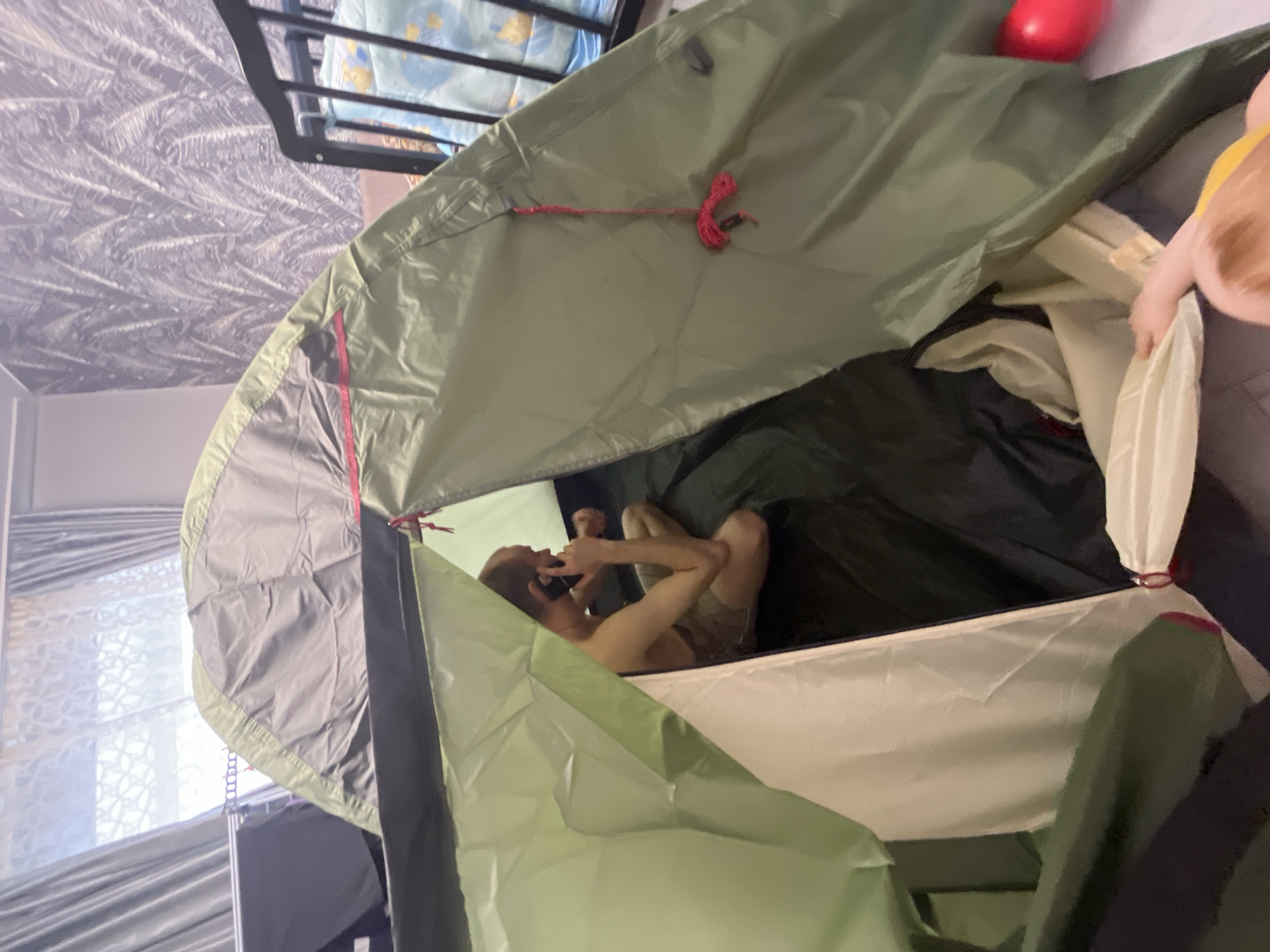 A man is inside a green and grey tent. He is lying down and holding his phone to his ear, appearing to be on a call. The tent is located indoors, with a bed frame and patterned wallpaper visible in the background. The location is Chaikovsky, Russia.