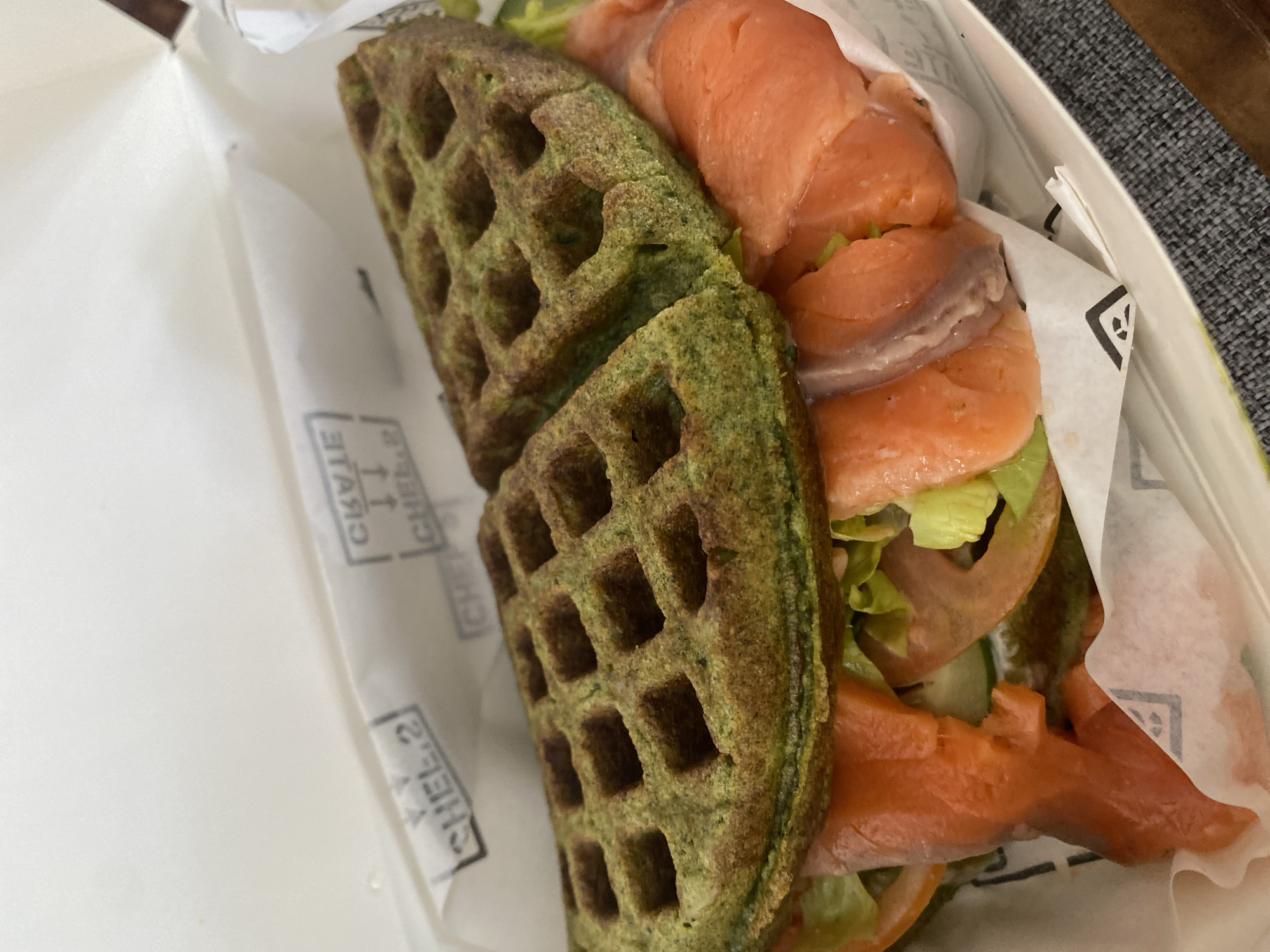 The image shows a green waffle sandwich, likely containing smoked salmon, lettuce, and tomato slices. The sandwich is in a white take-out container with printed branding, possibly "CHEF 2". The background is a simple, unadorned surface. There is no evidence to suggest the location is in Kuta Selatan, Indonesia. No people or interactions are visible.