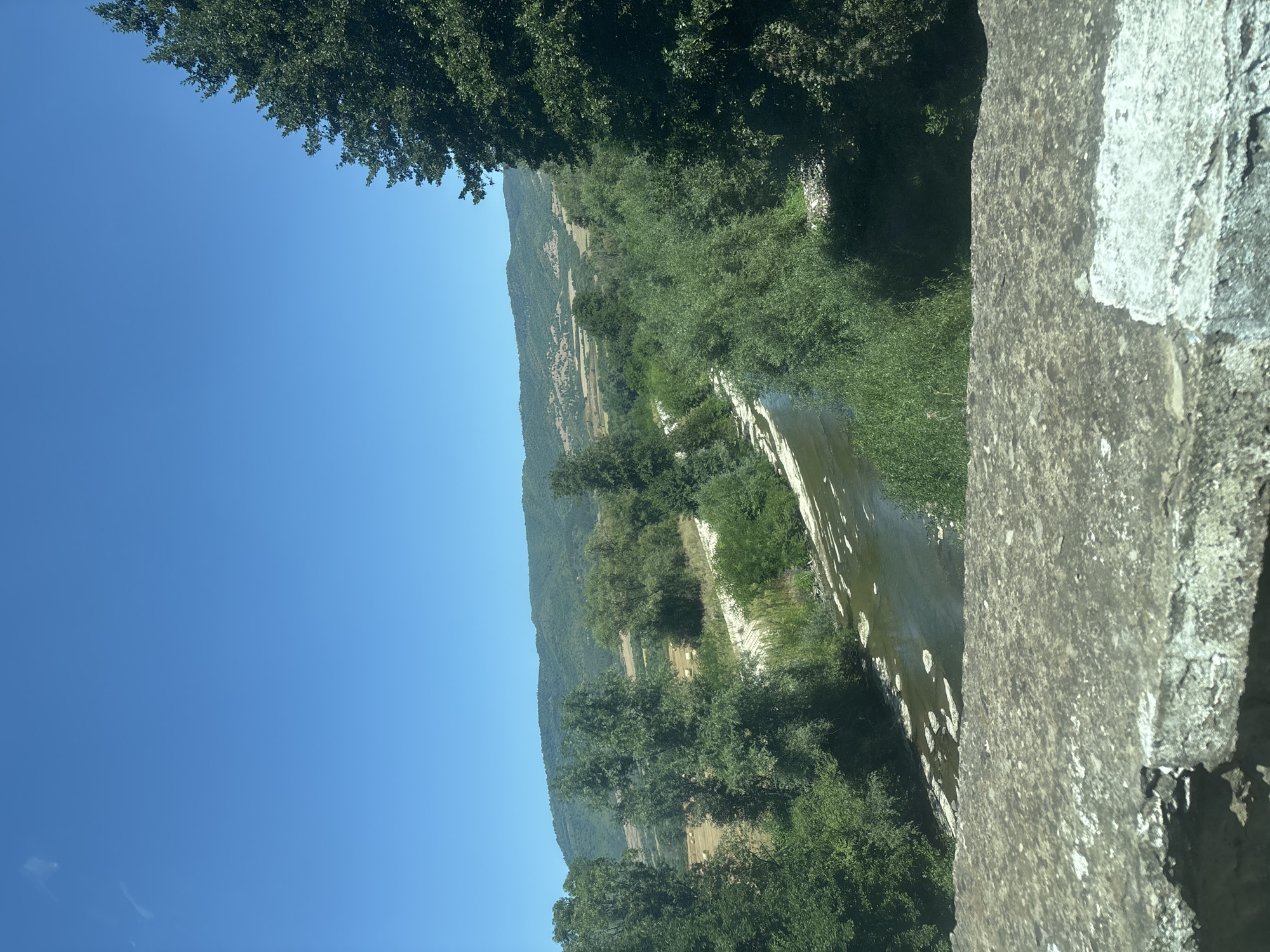 The image shows a river flowing through a lush green landscape under a clear blue sky. In the foreground, a rough concrete structure, possibly a bridge railing, is visible. The river is surrounded by trees and vegetation, with hills in the background. The water has a slightly murky appearance with some foam or debris on its surface. The overall scene depicts a natural environment, likely a rural or semi-rural area, on a bright sunny day. No people or specific city landmarks are identifiable in the image, and therefore, a specific location in Dzhurovo, Bulgaria cannot be confirmed.
