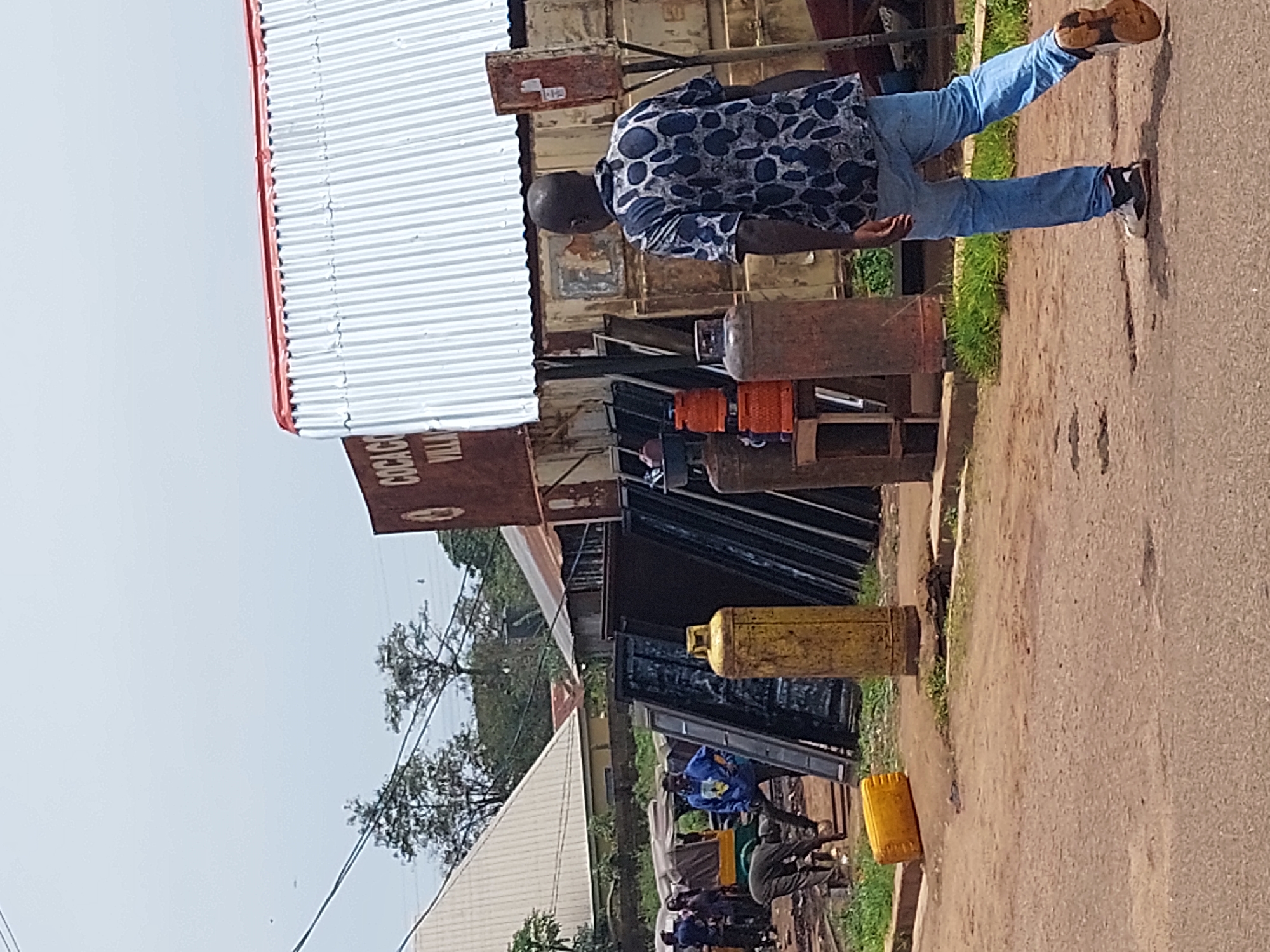 The image depicts a street scene in Barakin, Nigeria, on a bright, overcast day. The foreground is dominated by a dirt and asphalt road. On the right side of the road, a Black man wearing a blue and black patterned short-sleeved shirt and blue jeans is walking away from the camera, his right leg extended backward. He is wearing athletic shoes with white and black accents and brown soles.
Further back on the right, there are several large, cylindrical propane tanks. One is a dark reddish-brown color, and another is a bright yellow. Behind these tanks is a corrugated metal structure with a red trim roof, possibly a shop or workshop. A rusty, red metal box is mounted on a pole to the right of the entrance to the structure. A sign with "COCAGO VILLA" is visible on the front of the structure.
On the left side of the image, near the road, there is a yellow jerrycan lying on its side. A group of people are gathered in the middle ground, appearing to be working or engaged in some activity. Several large, flat, dark-colored objects, possibly metal sheets or doors, are leaning against the structure. A three-wheeled vehicle, a "keke napep," is parked near the group of people. The environment includes sparse green grass and trees in the background. Power lines are visible crossing the sky.