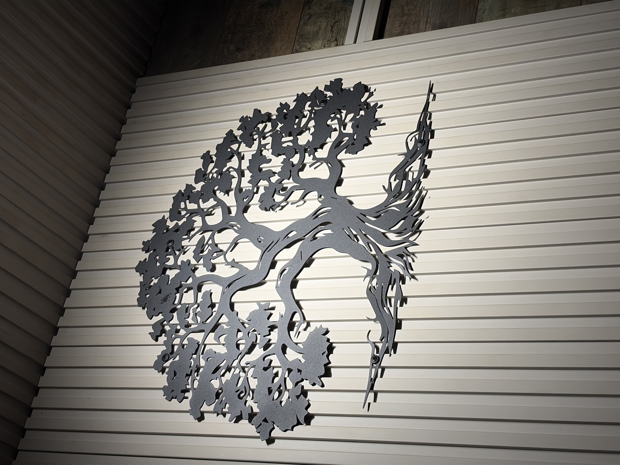A dark grey metal cutout of a stylized tree with leaves, branches, and roots, is mounted on a wall of horizontally oriented, narrow, white panels. The tree is positioned towards the center of the frame, with the left side appearing bushier with leaves and the right side appearing to transform into long, wispy strands, reminiscent of feathers or wings. A screw is visible fastening the metal cutout to the wall on the right. The wall appears to be part of an interior space, with another adjacent wall visible on the left, constructed of similar white panels oriented diagonally. The lighting suggests a single source from the left side, casting shadows and highlighting the texture of the wall. The location is not determinable from this image, as it lacks specific identifiers.
