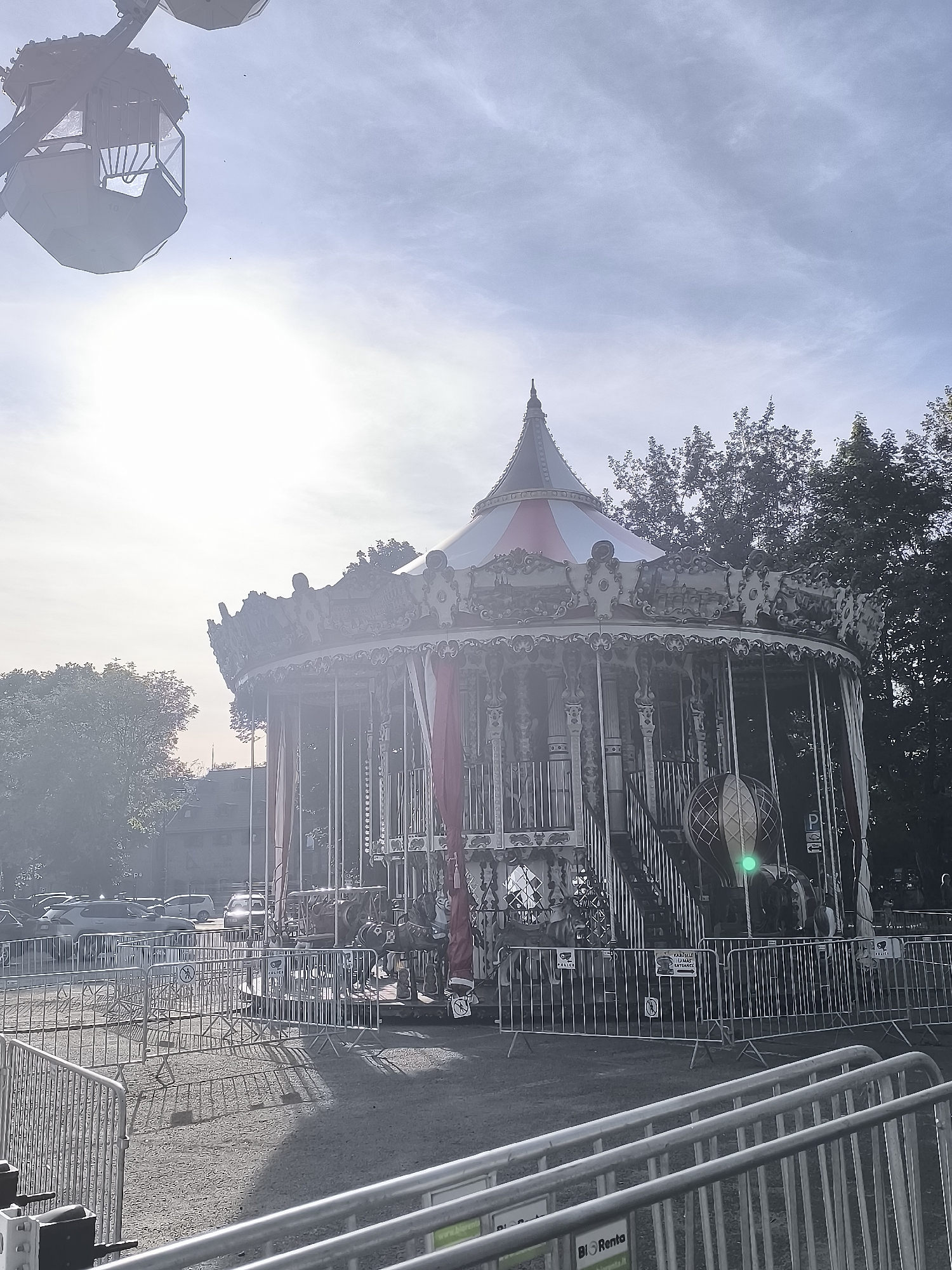 The image shows a carousel at an outdoor amusement park on a bright, hazy day. The carousel is ornate with decorative elements and red drapes hanging from its sides. In the background, trees and buildings are visible. A Ferris wheel gondola is also visible in the upper left corner of the image. The ground around the carousel is gravel, and metal barriers are set up around the perimeter. The scene appears to be in a public outdoor space, possibly a park or fairground.
