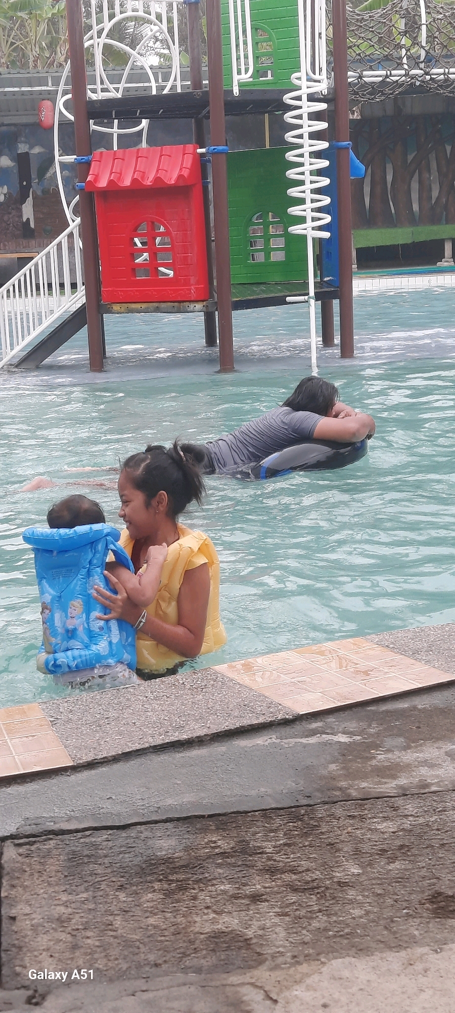 In the foreground, a young girl in a yellow life vest is holding a baby wearing a blue floatation device adorned with cartoon characters. They are in shallow water at the edge of a swimming pool, with a tiled pool border visible. In the middle ground, another person in a grey rash guard is floating on a black inner tube, resting their head on their arms. The background features a brightly colored children's playground structure with red and green sections, a white slide, and a coiled white spring. The environment appears to be an outdoor swimming pool area. The location is identified as Popoh, Indonesia, suggesting a recreational water park or similar facility.