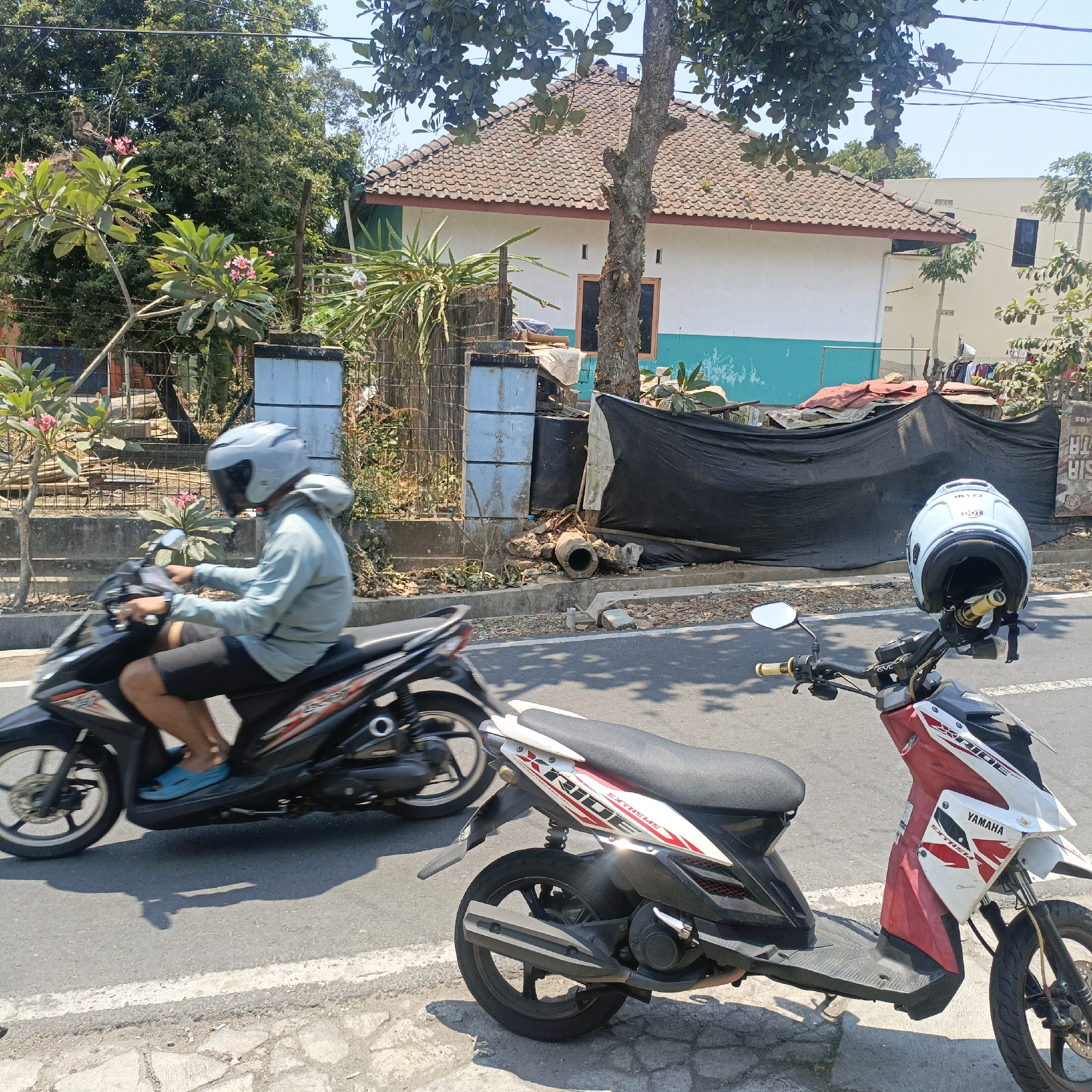 In Salatiga, Indonesia, a person wearing a light blue hooded jacket and helmet rides a black and white motorcycle. Another red and white motorcycle is parked in the foreground, with a light blue helmet resting on its handlebars. The motorcycles are on an asphalt road with a stone-lined curb. Behind the road, a fence encloses an area with a simple house featuring a terracotta tile roof and white and turquoise walls. Trees and foliage are visible, and some debris and construction materials are piled near the fence. The sky is clear and blue, suggesting a sunny day.