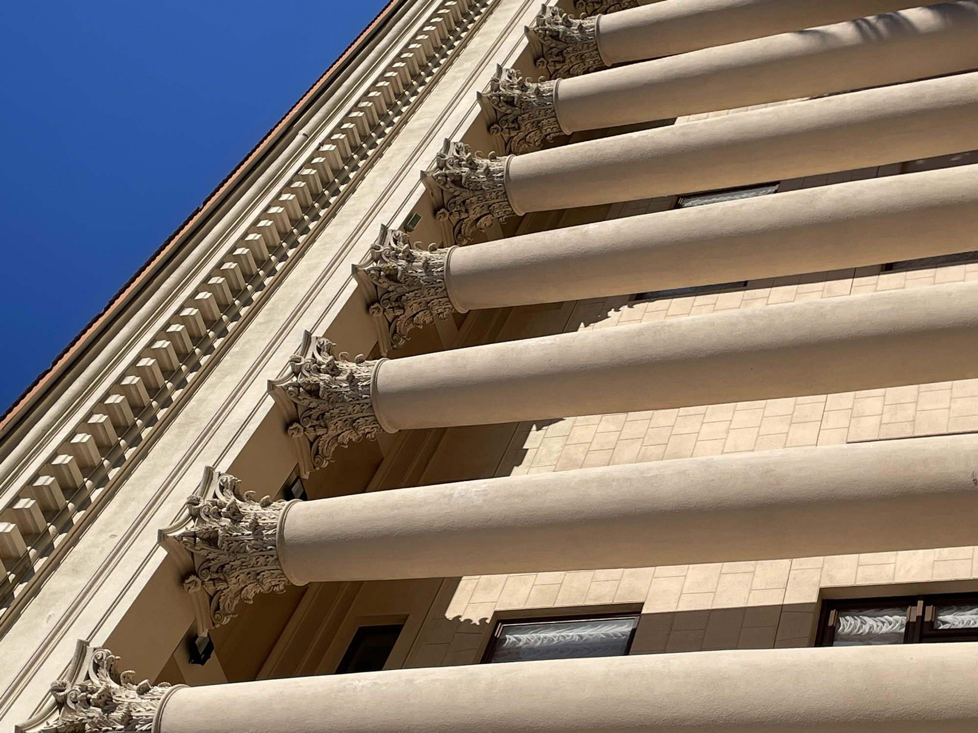 The image captures a close-up, low-angle view of a building facade featuring a series of large, ornate Corinthian columns made of light-colored material. The columns are closely spaced and extend upwards, supporting an architectural entablature with decorative molding and a patterned frieze. Above the entablature, the roofline is visible, capped by a red tile roof, and a clear, deep blue sky fills the upper right portion of the frame. The building wall behind the columns is constructed of light beige brickwork, with dark-framed windows visible between the columns. The sunlight casts strong shadows, highlighting the intricate details of the column capitals and the architectural elements of the building. This building appears to be a public or institutional structure, possibly a government building, museum, or theater, given its classical architectural style. Based on its architectural style and grandeur, this could be the Winter Theater in Sochi, Russia. There are no people or animals visible in the image, and no discernible action is taking place.
