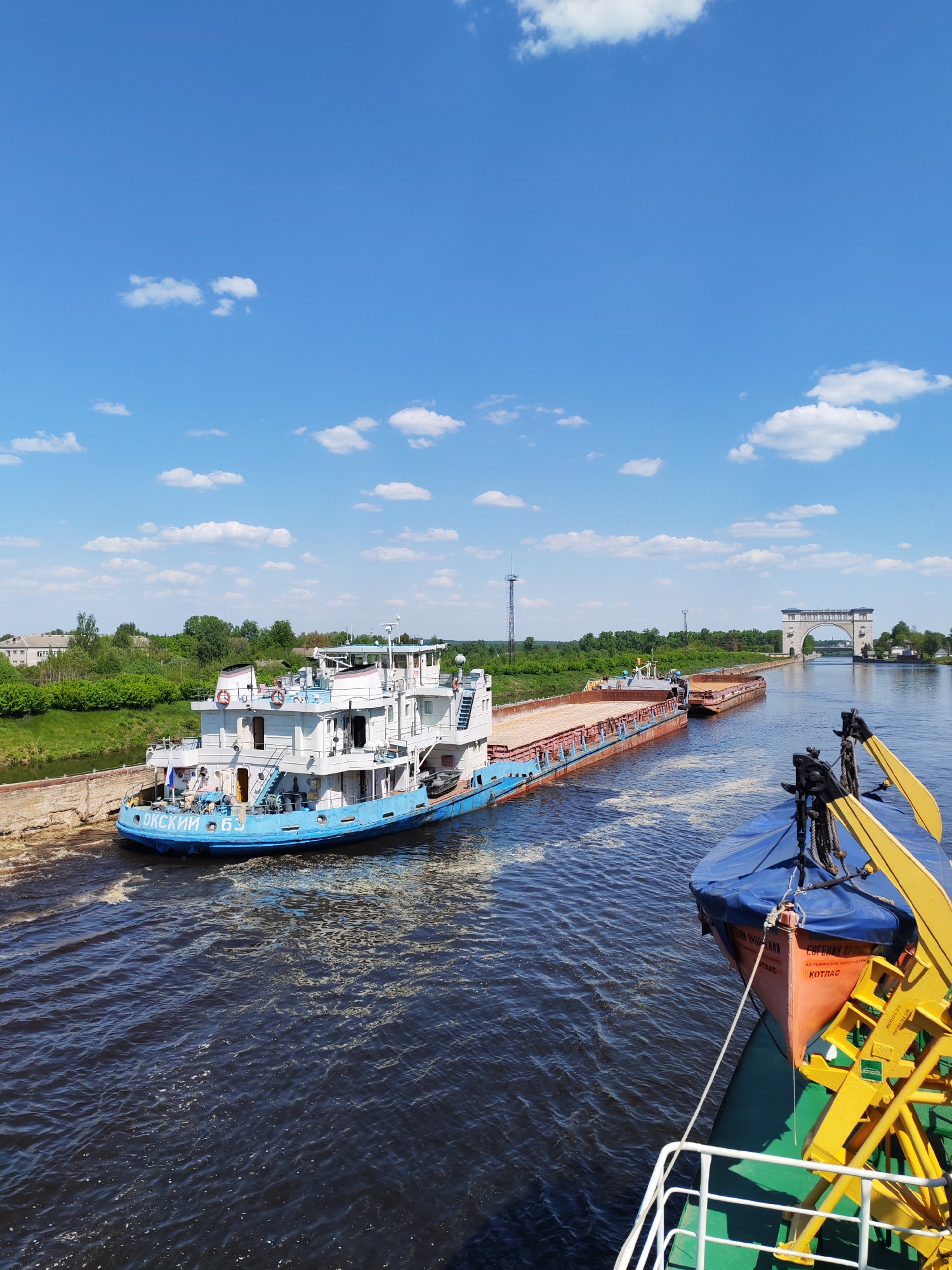 A photograph captures a waterway scene under a partly cloudy sky. A blue and white boat is moored near a concrete bank to the left. The boat has Cyrillic lettering in blue on its hull. Connected to the boat is a series of barges, visible further down the waterway. A tall, arched structure spans the river in the distance.
The foreground features a portion of another vessel, with yellow and blue structures. An orange lifeboat is visible on the side of the vessel, and Cyrillic text is present on the boat.
The water is dark and slightly turbulent. The banks are green with trees and vegetation, and a few buildings are visible in the background. There are utility masts and overhead electrical infrastructure visible to the left and further down the waterway.