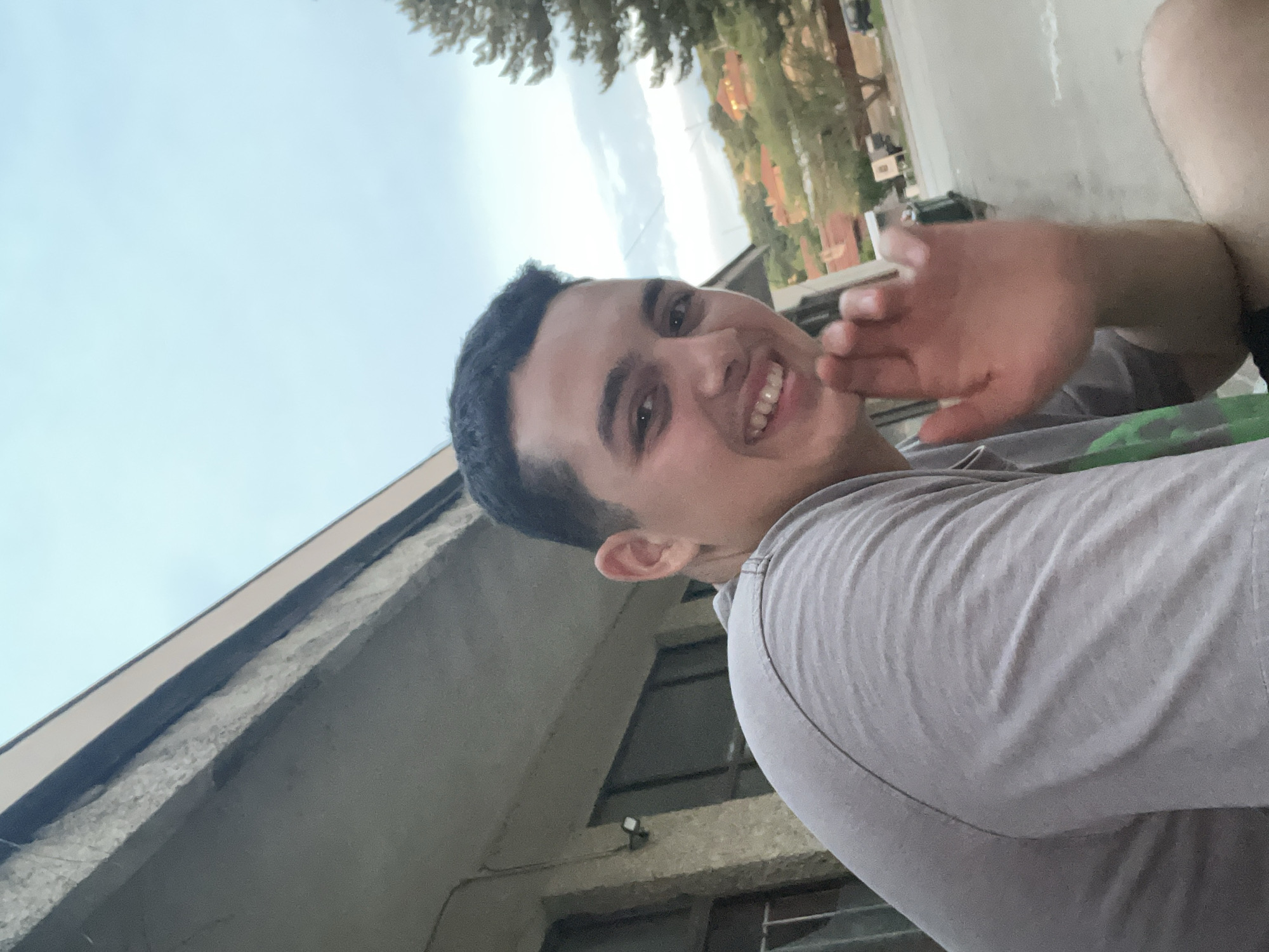 A male individual, appearing to be a young adult, with short dark hair and a light complexion, is depicted in a chest-up, outdoor shot in the city of Basarbovo, Bulgaria. He is positioned on the right side of the frame, smiling broadly with his teeth visible, and looking directly at the camera. His right hand is raised, palm facing forward, in a gesture resembling a wave. He wears a light grey short-sleeved t-shirt, with a hint of a green pattern visible on the lower left side of his torso. The background on the left features a portion of a building with a textured, possibly concrete or stone, facade, dark rectangular windows, and a mounted outdoor light fixture. An exposed, rough architectural element is visible along the building's edge. To the right and in the distance, a road or street is visible, with trees and residential buildings situated on a sloped terrain. The sky above is light blue with scattered clouds.