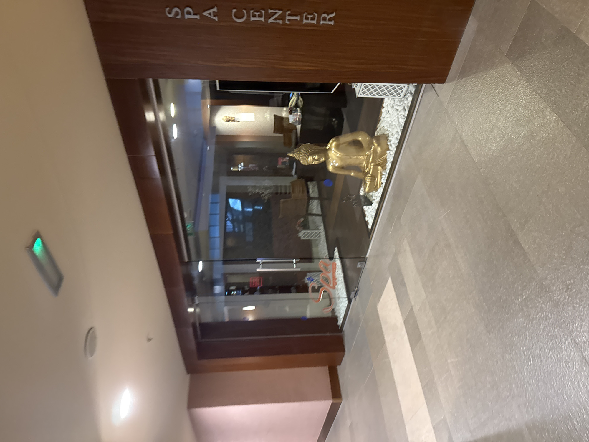The image shows the entrance to a spa center in Aksu, Turkey. The entrance is a glass door with wooden trim. Inside the spa, there is a golden Buddha statue sitting on a bed of white stones. The floor is tiled with a pattern of light and dark gray tiles. The walls are painted a light pink color. There is a sign on the wooden trim that says "SPA CENTER" in white lettering. The overall impression is one of tranquility and relaxation.