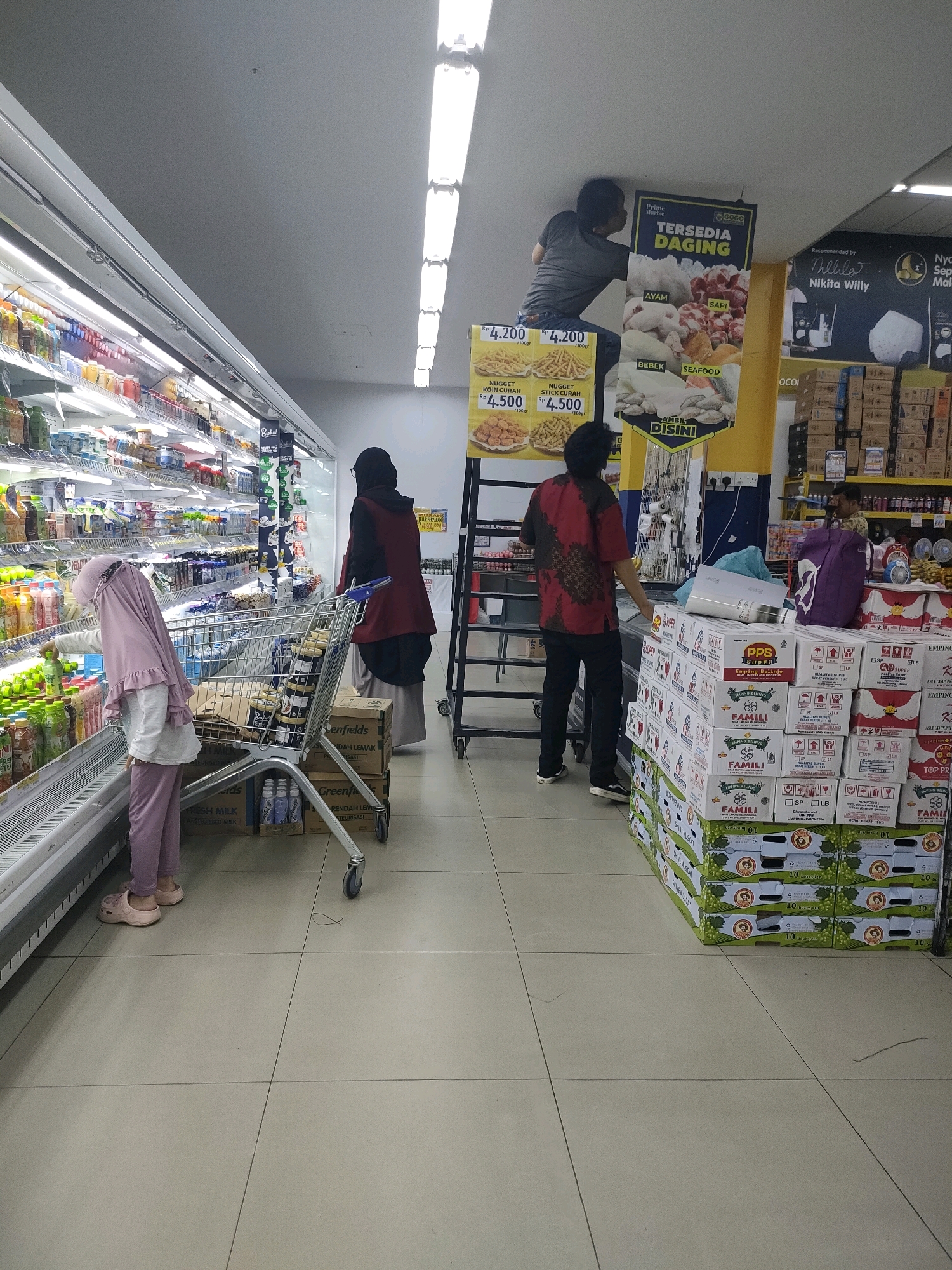The image depicts the interior of a supermarket, likely located in Batam, Indonesia. In the foreground, a person wearing a pink headscarf and pink pants is looking at products in a refrigerated display case. To their right, a shopping cart is positioned. Further back, another person in a dark headscarf and maroon outer garment stands near the shopping cart.
In the background, one person is standing on a ladder, appearing to be working on the ceiling or a high shelf. Another person in a red and black patterned shirt is looking at boxes stacked on the floor. The supermarket is well-lit with overhead fluorescent lights. Shelves are filled with various food and beverage products. A large banner displaying images of meats with text such as "TERSEDIA DAGING" (Meat Available), "AYAM" (Chicken), "SAPI" (Beef), "BEBEK" (Duck), and "SEAFOOD" is prominently displayed. Another banner with a portrait and text mentioning "Nikita Willy" is visible. Several stacks of boxes, some with the brand name "FAMILI," are also present. The flooring is tiled. The overall environment suggests a commercial retail space.