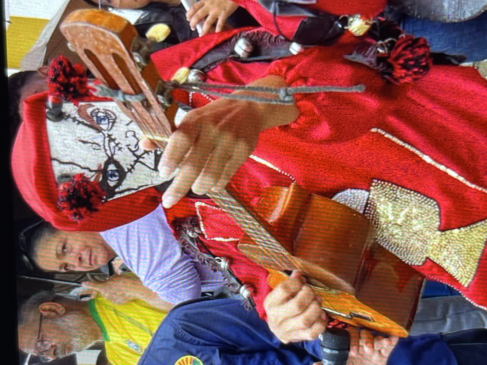 A person in a red costume with a mask depicting a skull-like face is playing a ukulele. The mask has blue eyes and black lines resembling cracks. The costume is adorned with gold embellishments and red pom-poms. The person is holding the ukulele with their left hand on the neck and their right hand strumming the strings. In the background, other people are visible, some holding umbrellas, suggesting an outdoor event. One person in the background is wearing a yellow shirt with green and blue accents, possibly a national flag design. The overall scene appears to be a public gathering or festival in Tinaquillo, Venezuela, characterized by traditional attire and musical performance.