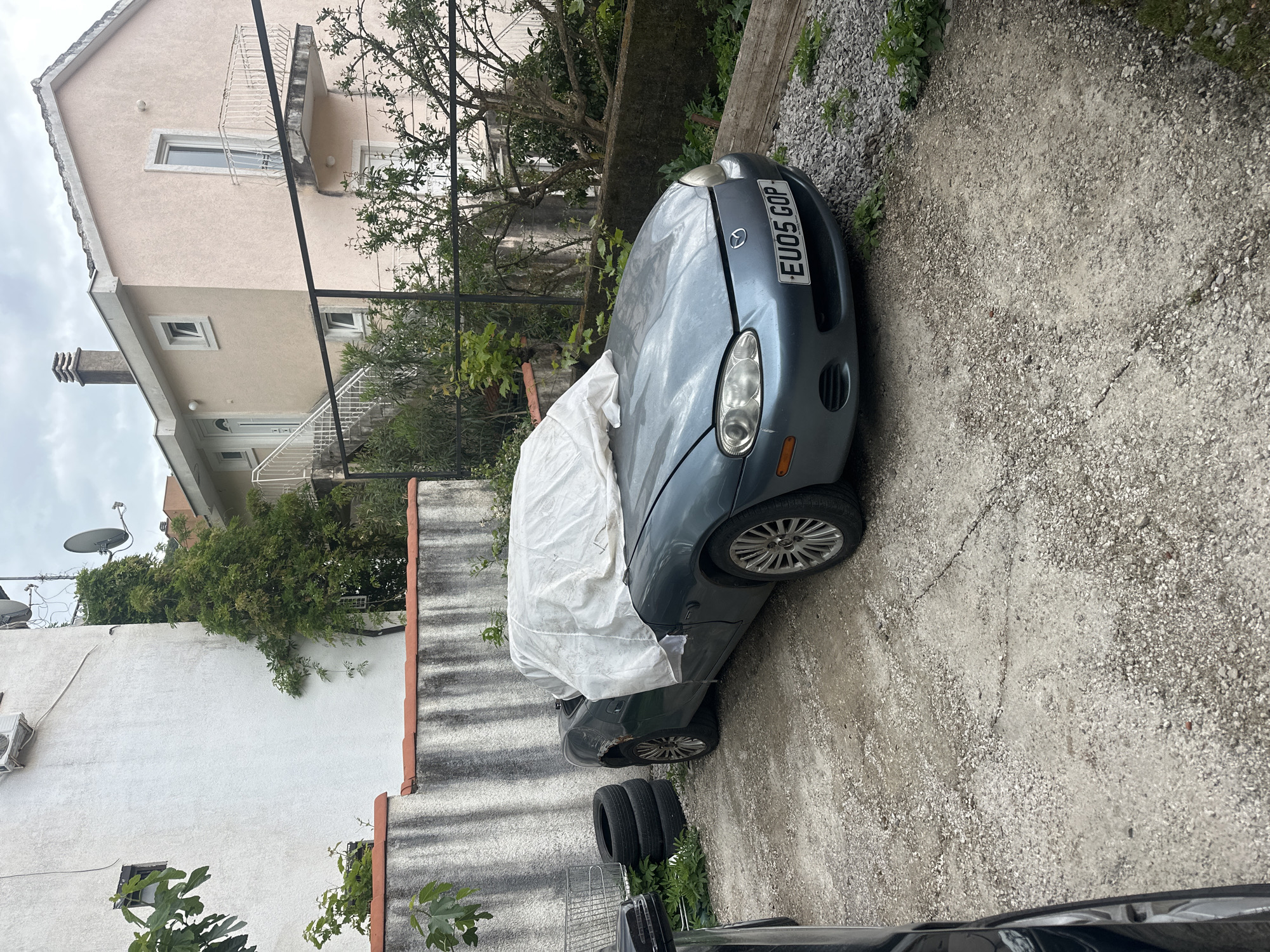 A light blue Mazda MX-5 convertible with UK license plate "EU05 GOP" is parked in Budva, Montenegro. The car is partially covered with a white sheet. It is parked on a gravel driveway next to a concrete wall. A house is visible in the background. Three tires are visible on the ground next to the concrete wall. Another car is partially visible in the lower right corner of the image.