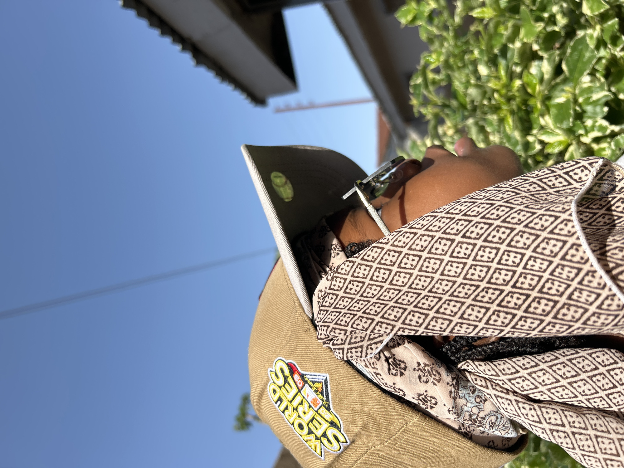 A low-angle shot depicts a person wearing a beige baseball cap with a "World Series" patch, sunglasses, and a patterned shirt or scarf. The person's face is partially visible, and their hair appears dark. The background includes a clear blue sky, a building, and some green foliage. There is not enough information to identify the location as Jos, Nigeria.