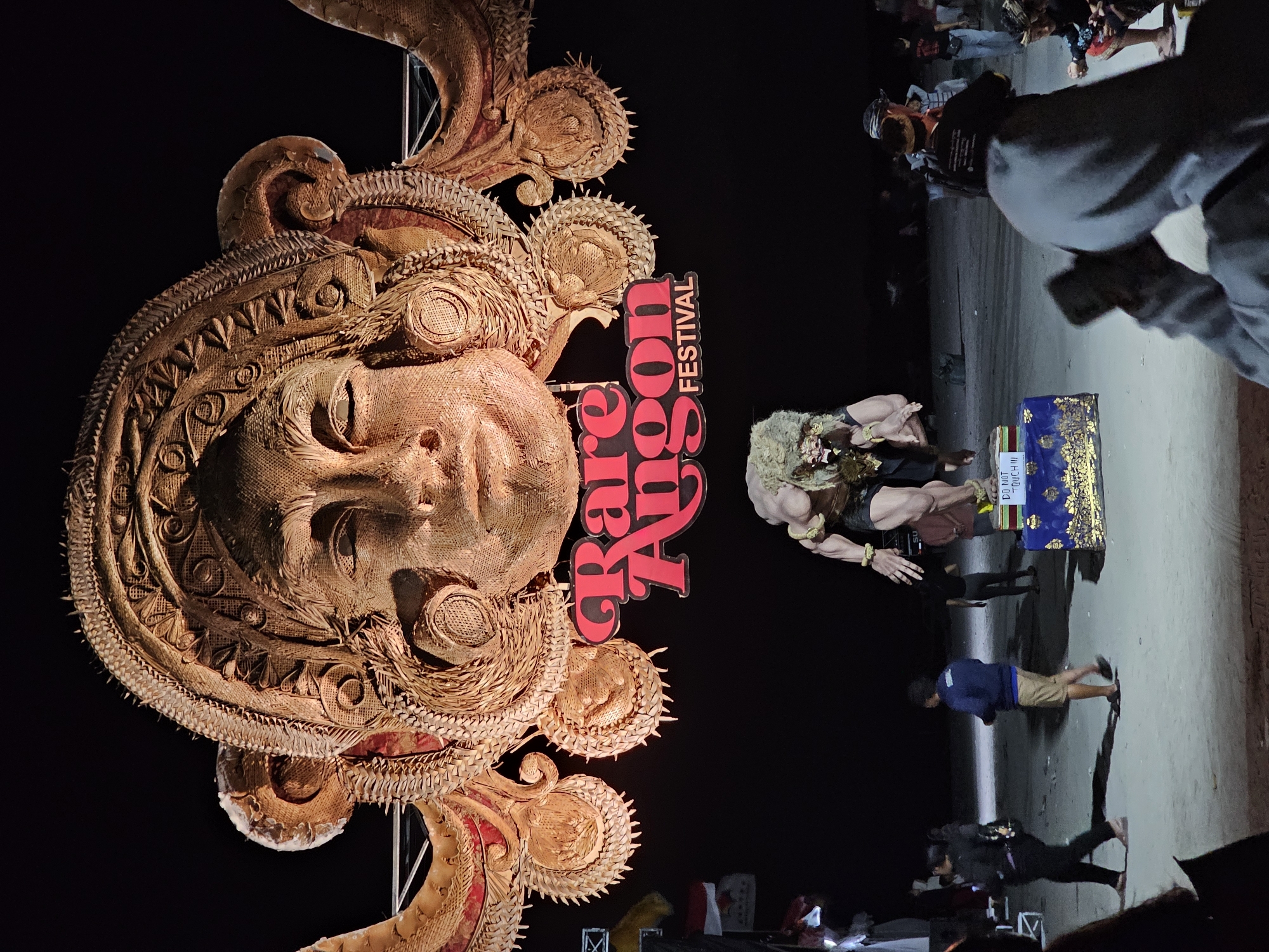 The image shows a large, intricately crafted effigy of a face with elaborate headdress, likely a Rakshasa or demonic figure, prominently displayed at night. Beneath the effigy, a smaller figure of a muscular, creature-like being stands on a decorated base with a "DO NOT TOUCH!!!" sign. The scene appears to be outdoors on a sandy area, possibly a beach, with a dark sky and artificial lighting illuminating the figures and the event signage, which reads "Rare Angon FESTIVAL" in large, red, stylized lettering. Several people are visible in the background and foreground, some walking, others observing the display. The overall atmosphere suggests a cultural festival or performance in Denpasar, Indonesia, given the context of "Rare Angon" which is a traditional Balinese art form and festival originating from Denpasar.