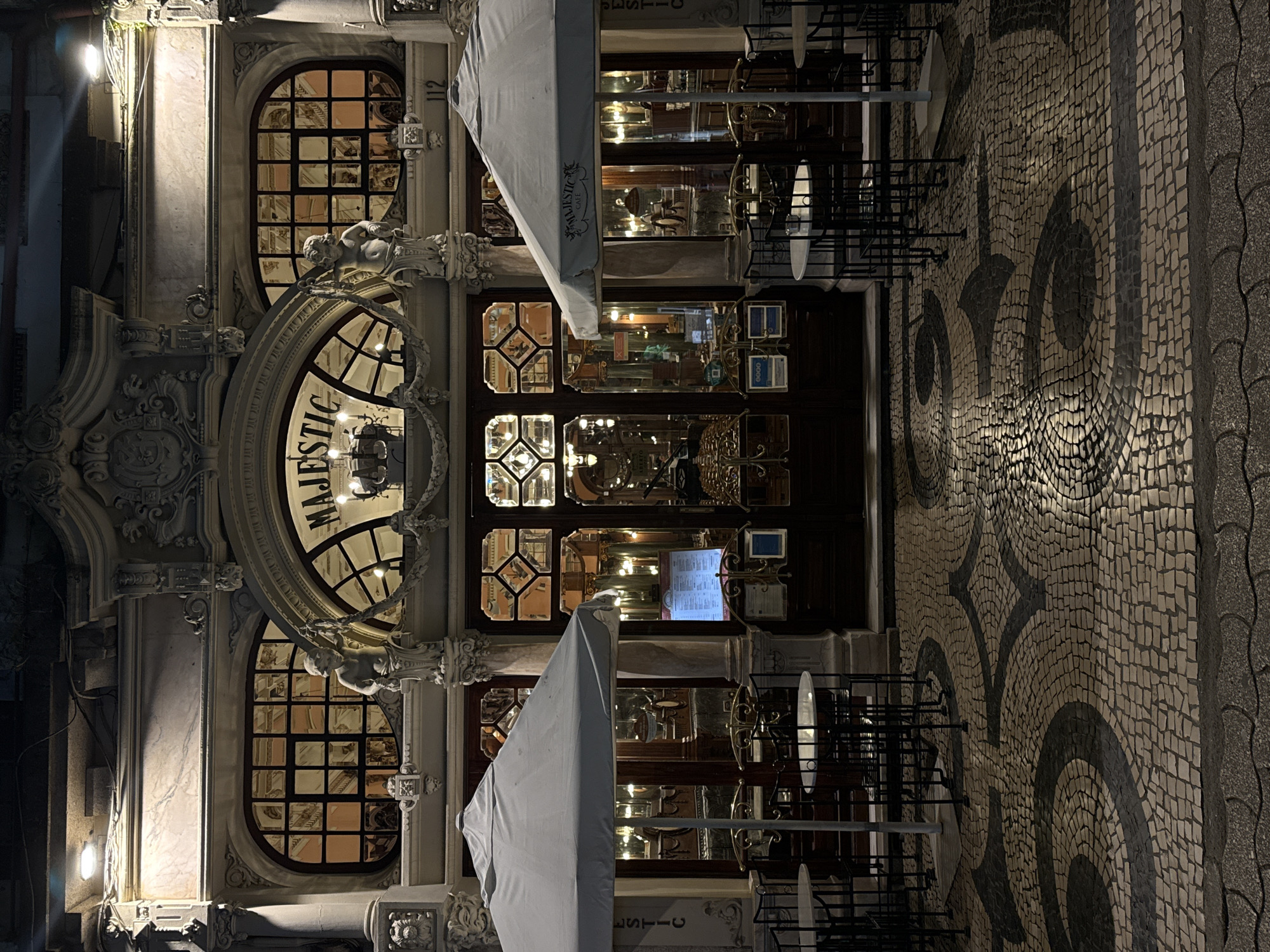 The image displays the illuminated facade of the Majestic Café, located at Rua de Santa Catarina 112, in the city of Porto, Portugal. The scene is captured at night.
The building's ornate Art Nouveau-style facade is constructed from a light-colored material, likely stone or stucco, featuring intricate carvings, columns, and decorative pilasters. The upper level includes a prominent central arched window displaying the word "MAJESTIC" in script, flanked by two cherub-like sculptures holding decorative garlands. Two oval-shaped windows with multi-pane grids are situated to either side of the central arch. The lower level consists of large display windows with dark wooden frames and intricate glass patterns, revealing the brightly lit interior of the café. The address number "112" is affixed above the lower right window.
In front of the café, an outdoor seating area is set up. Two large, light-colored patio umbrellas, each emblazoned with "MAJESTIC CAFE", are open and positioned on either side of the central entrance. Beneath each umbrella are multiple small, round tables with light-colored tops and dark metal bases, surrounded by dark metal chairs featuring ornate backs. A backlit menu stand is visible to the left of the main double-door entrance.
The ground directly in front of the café is paved with traditional Portuguese mosaic cobblestones (Calçada Portuguesa), arranged in a decorative black and light-colored curvilinear pattern. A darker, wavy-patterned cobblestone street or curb is visible at the bottom edge of the image.
No individuals are present in the scene, and no specific actions or interactions are depicted. The café appears to be operational, indicated by the interior lighting and displayed menu.